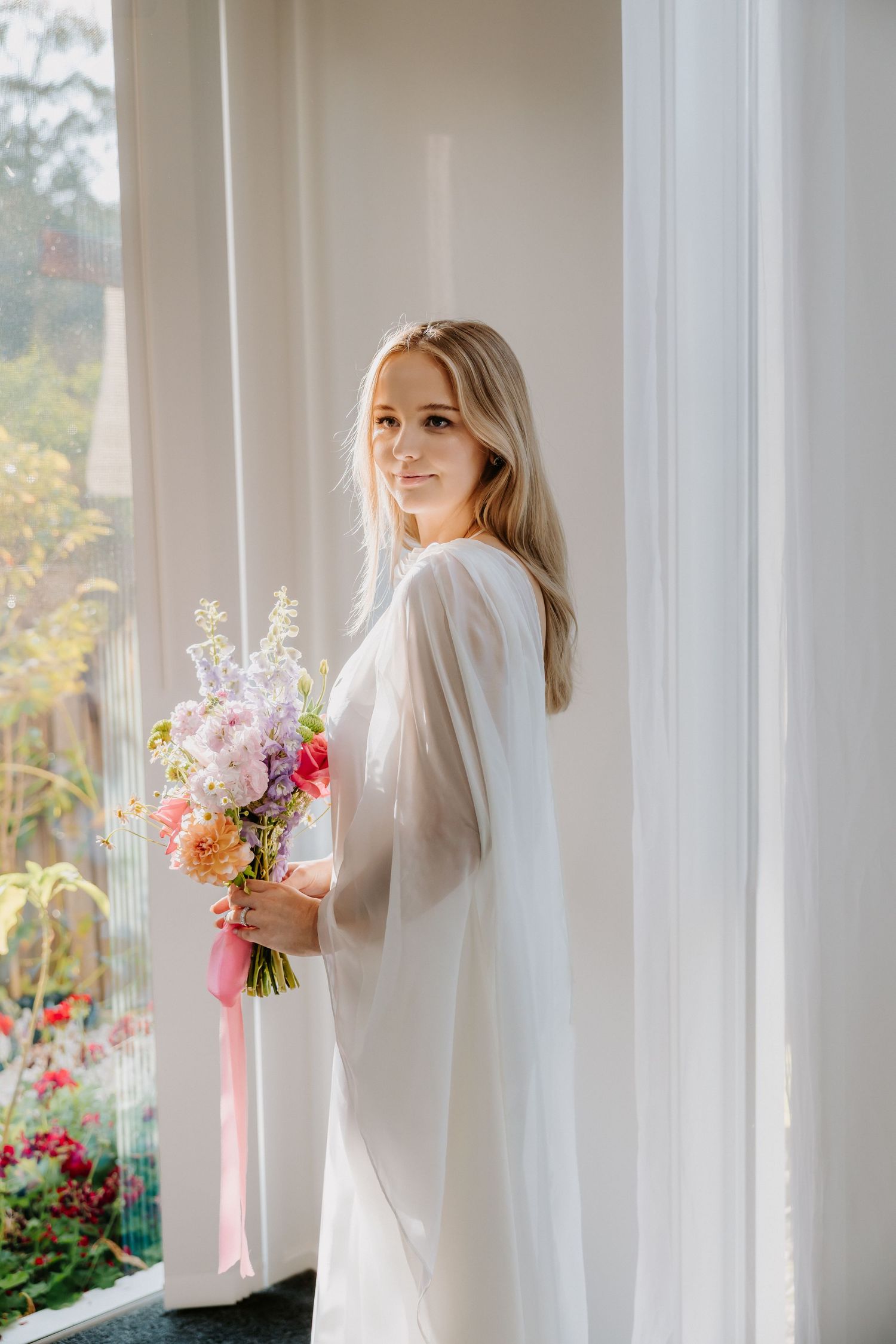 A person in white holding colorful floral bouquets near sunlit windows.