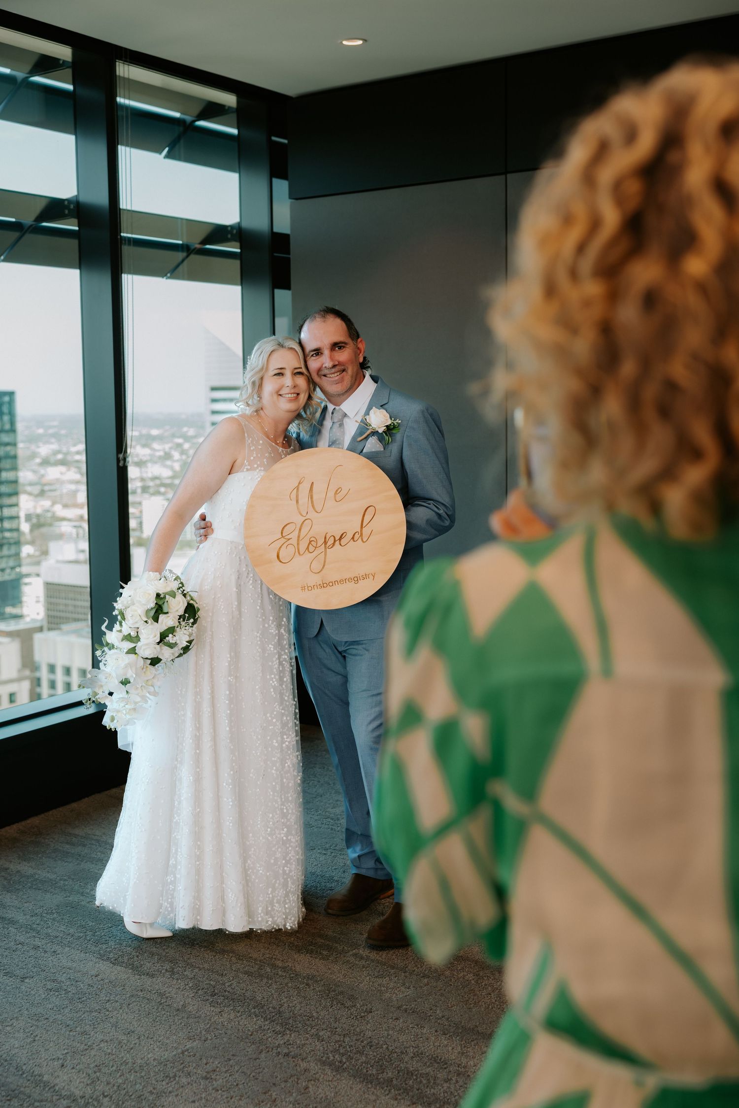 A couple poses for wedding photos in a modern indoor venue with floor-to-ceiling windows and city views.
