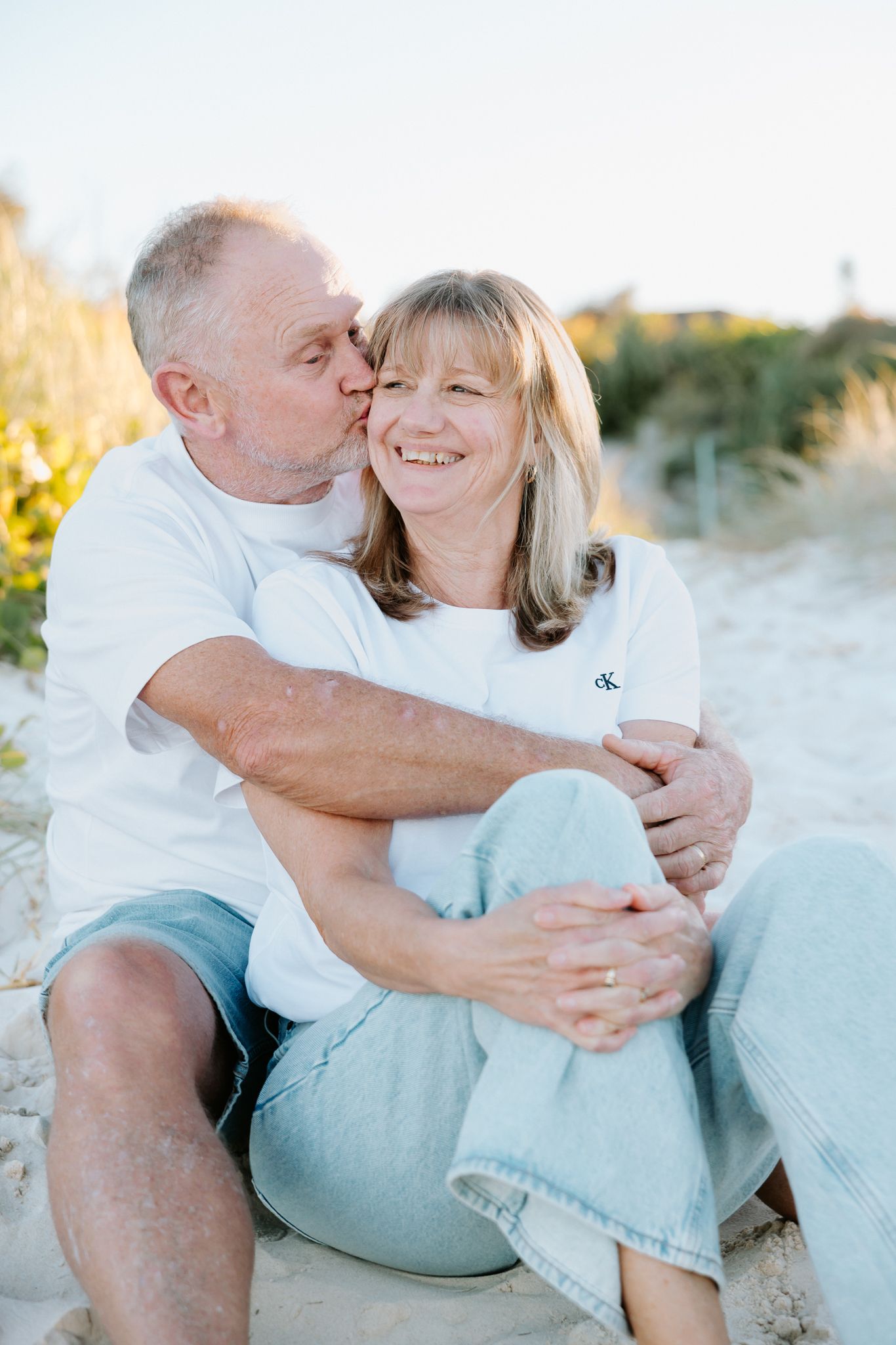 Middle-aged couple sitting on beach sand, man kissing woman on cheek, both wearing white t-shirts and light jeans.