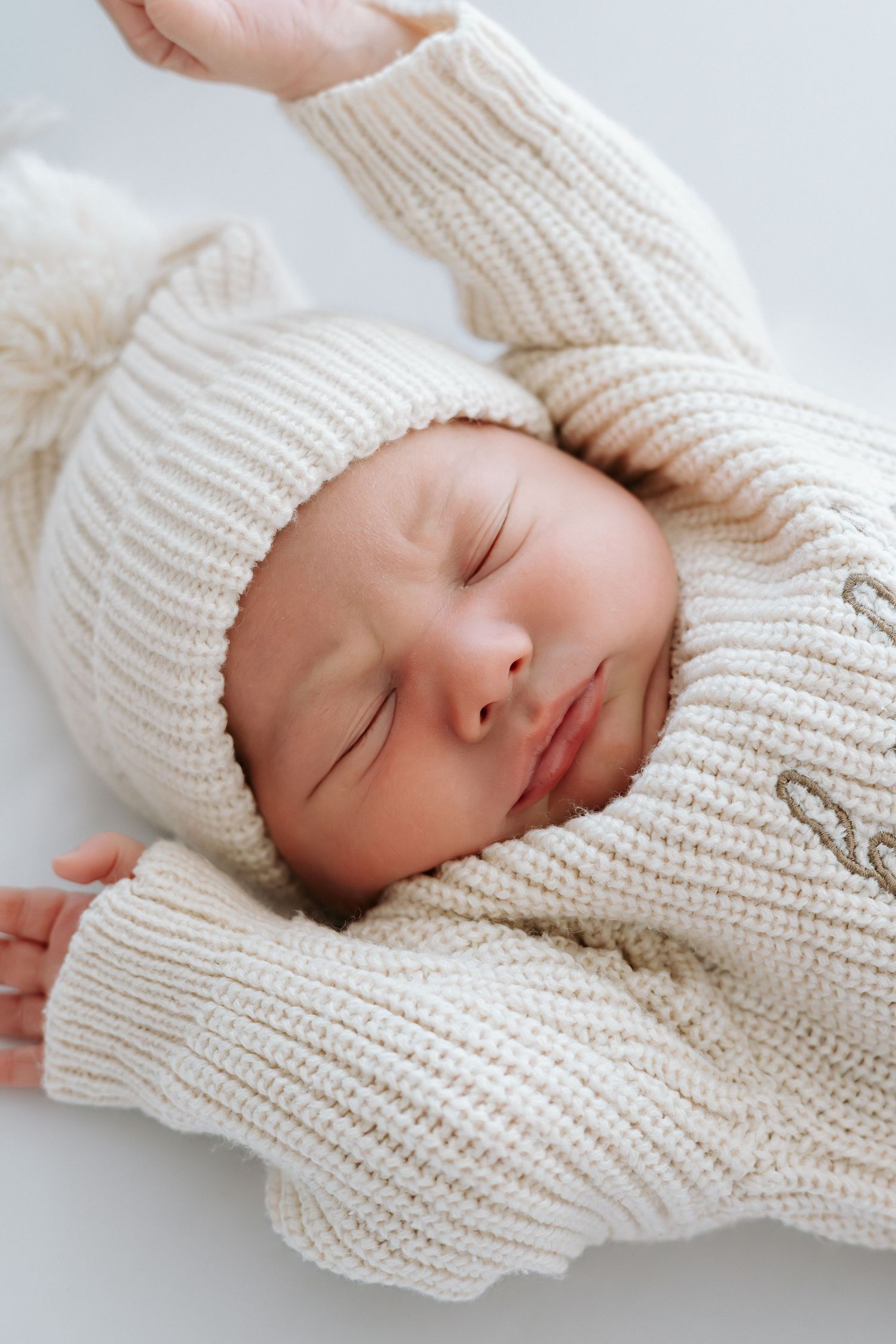 Peaceful newborn baby sleeping in cream knit sweater and matching pom-pom hat on white background.
