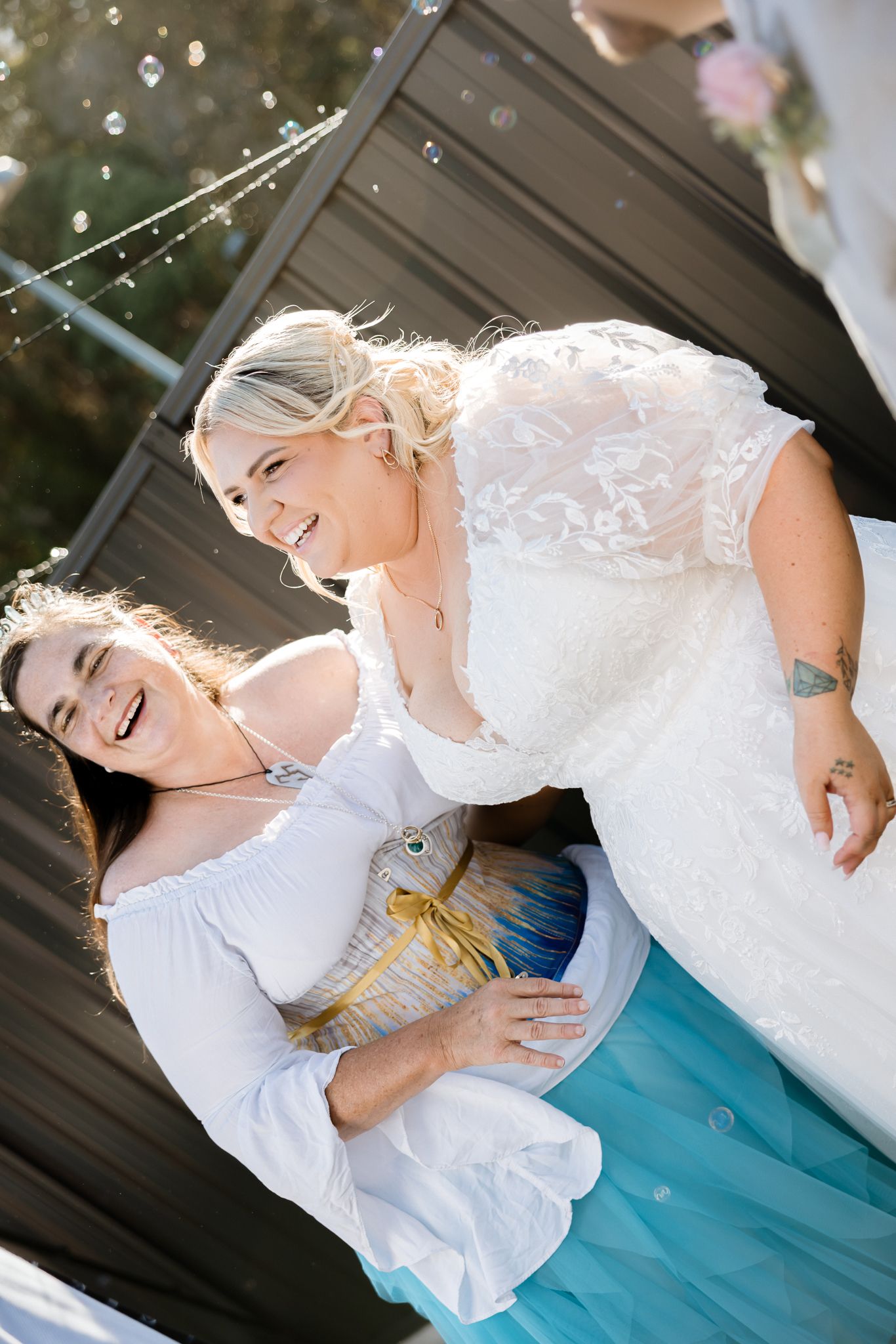Two women in white dresses laugh joyfully outdoors, one in a lace bridal gown, surrounded by bubbles at a wedding.