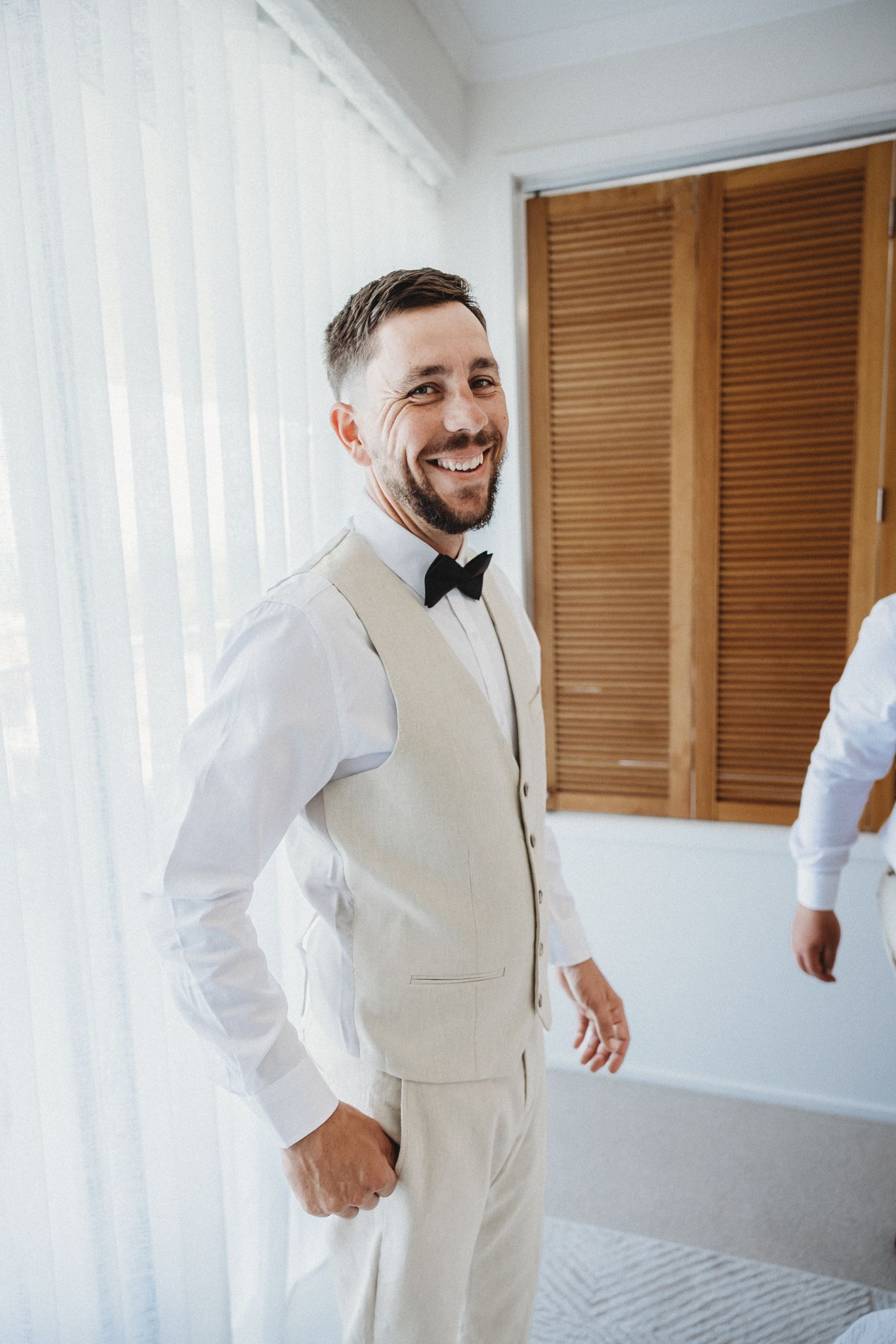 Smiling groom in cream vest and black bow tie posing indoors on wedding day.
