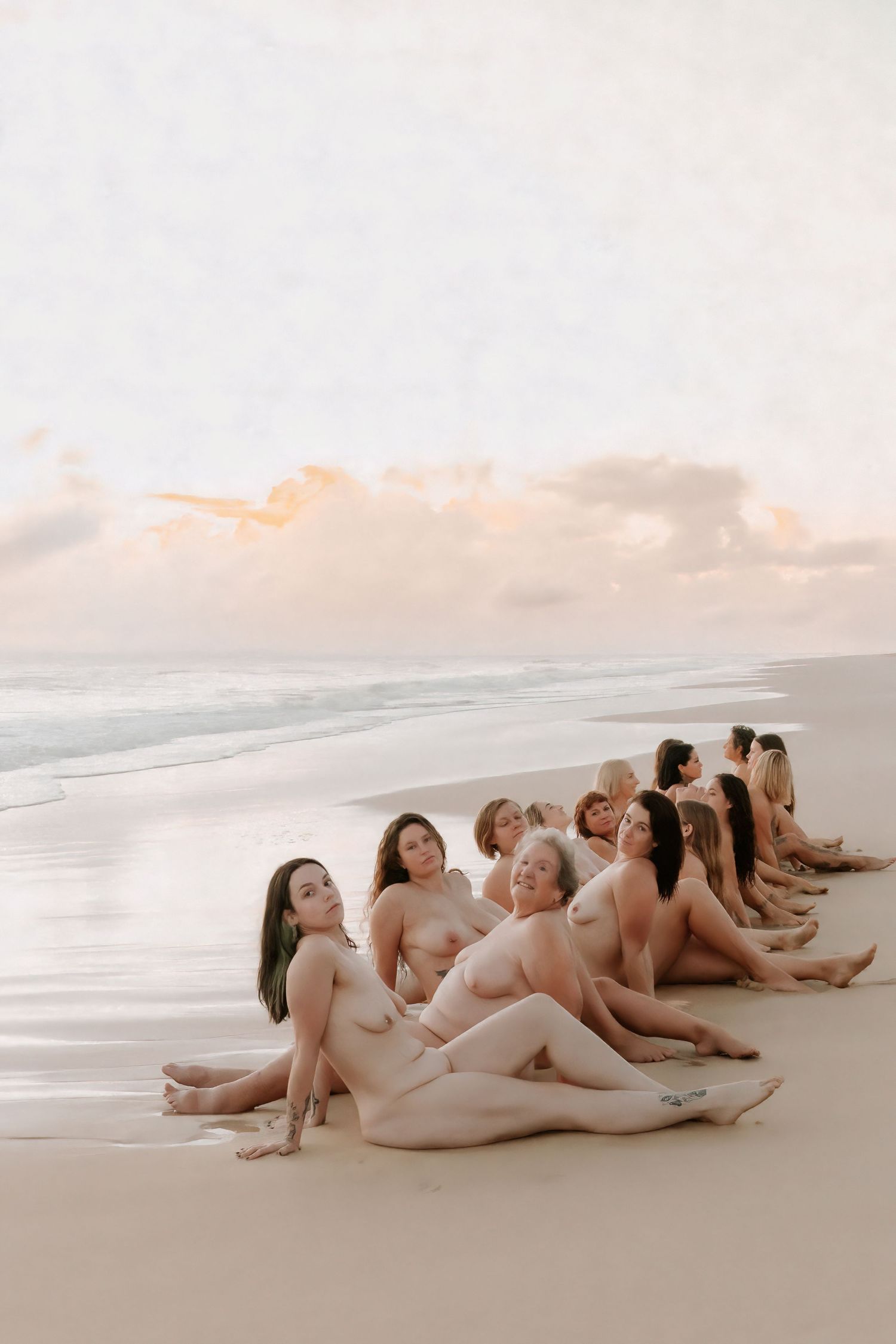 A group of nude women sitting together on a sandy beach at sunset with ocean waves in the background.