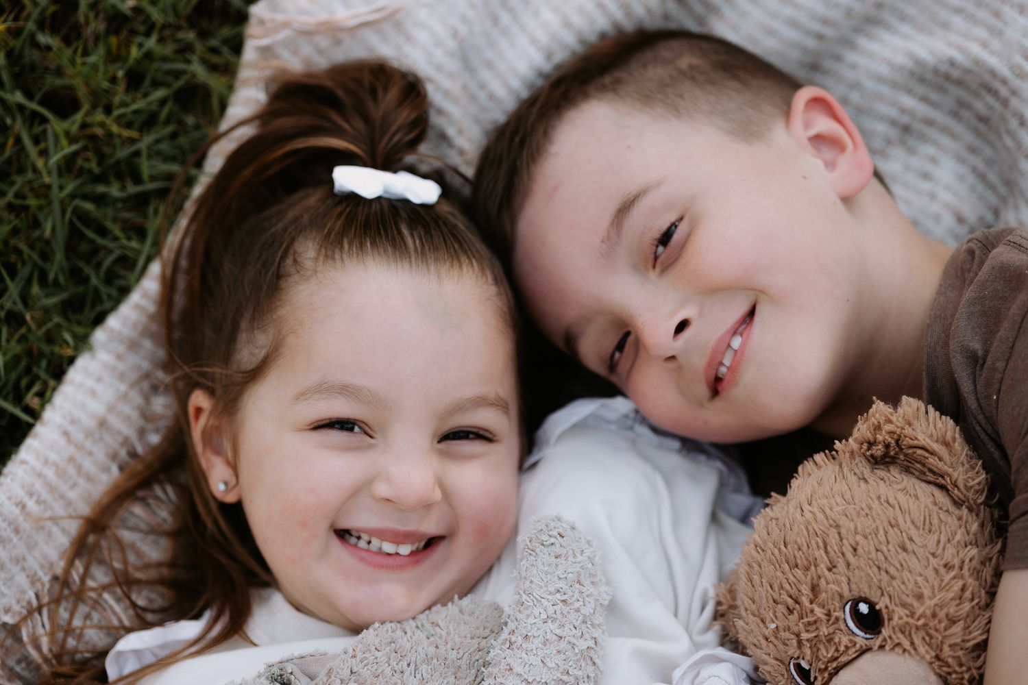 Two smiling children lying on a blanket outdoors, with a girl in a hair bow and a boy holding a teddy bear.