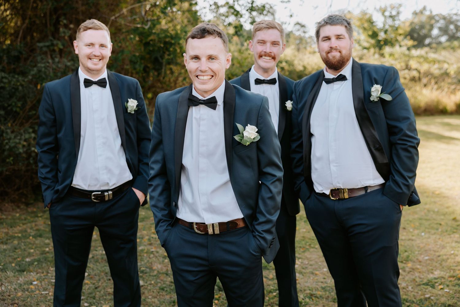 Four groomsmen in black tuxedos with bow ties and white boutonnieres posing outdoors at a wedding.