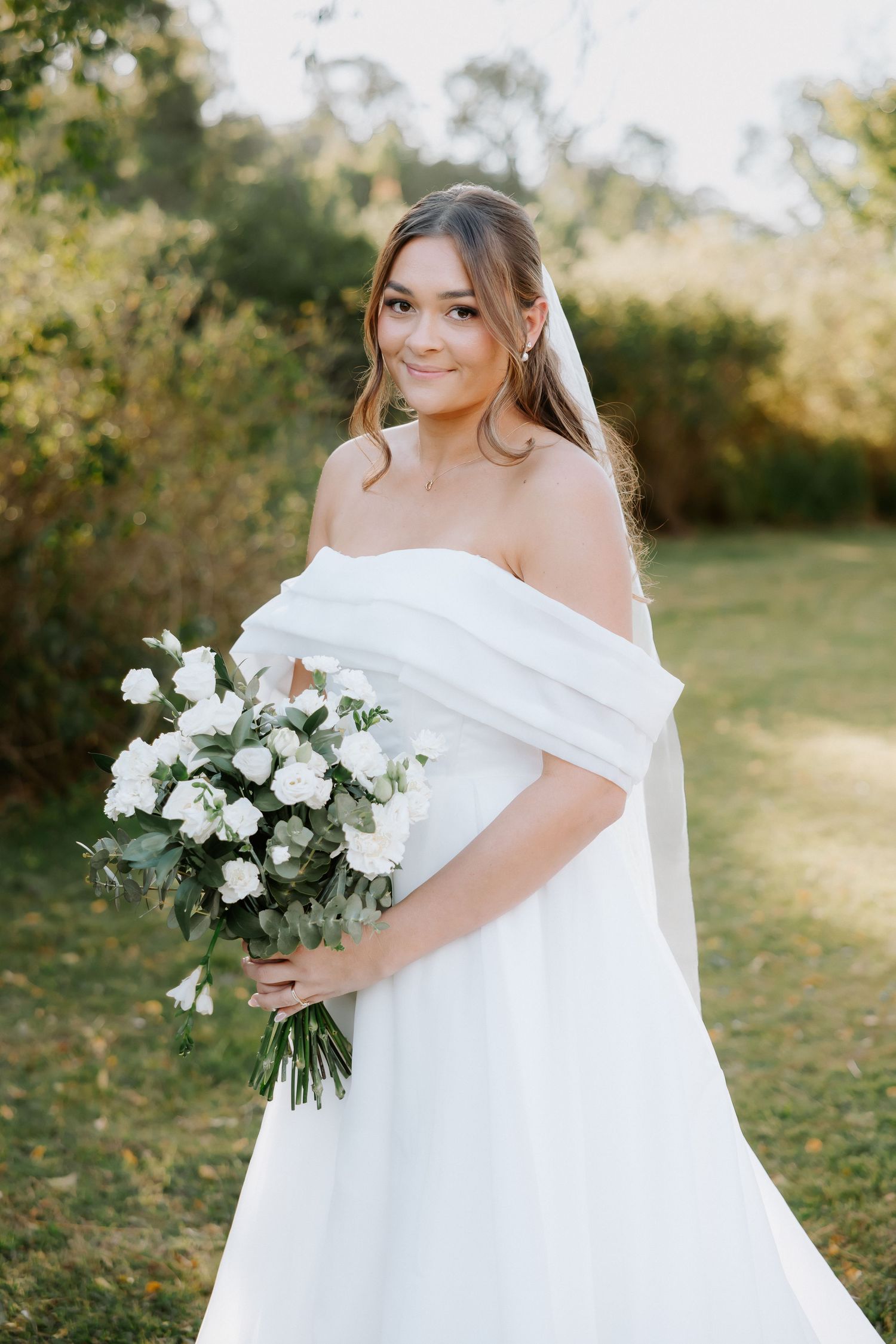 Bride in white off-shoulder gown holding white rose bouquet, smiling outdoors with green foliage background.