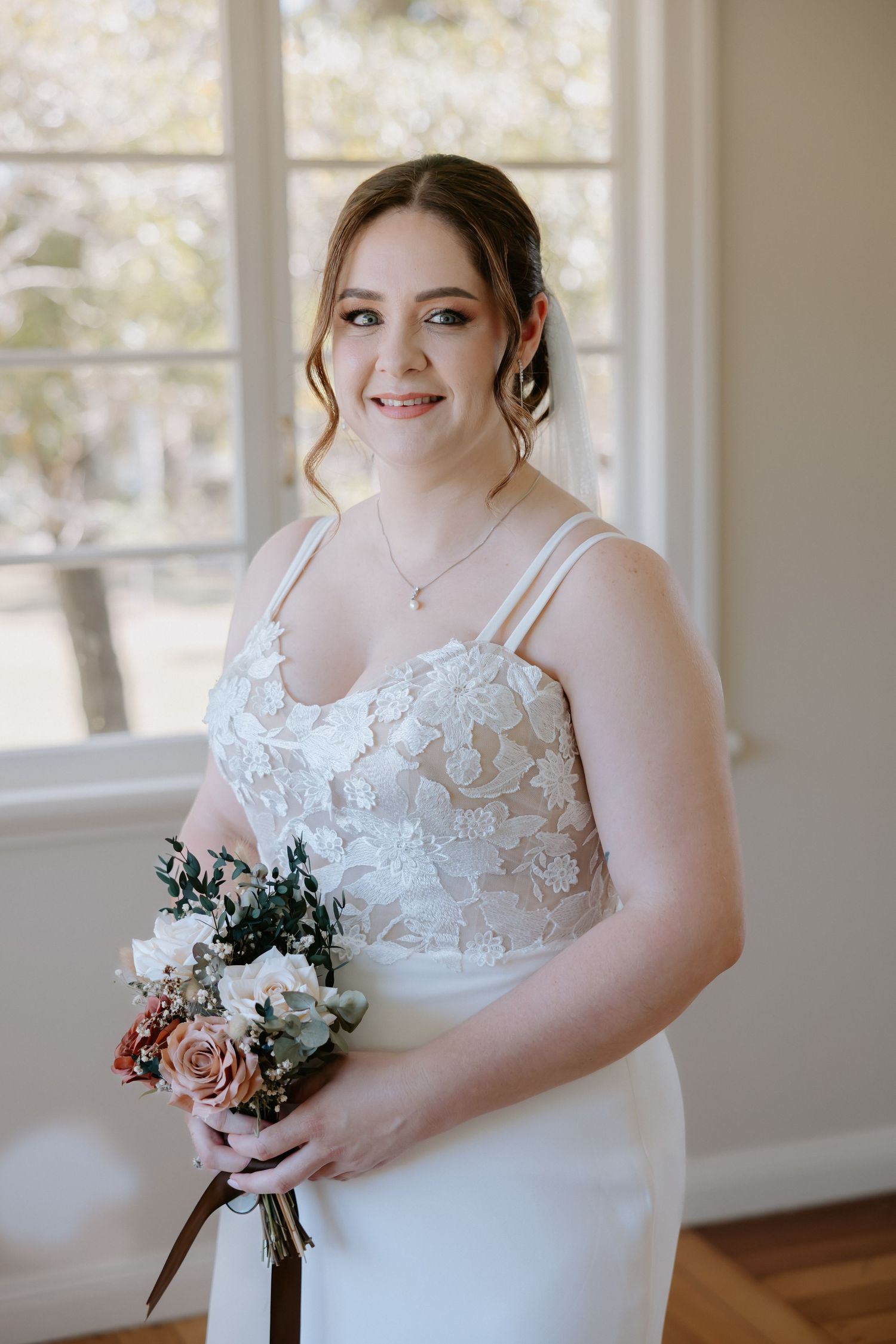 Bride holding a bouquet in a sunlit room with hardwood floors and white walls.