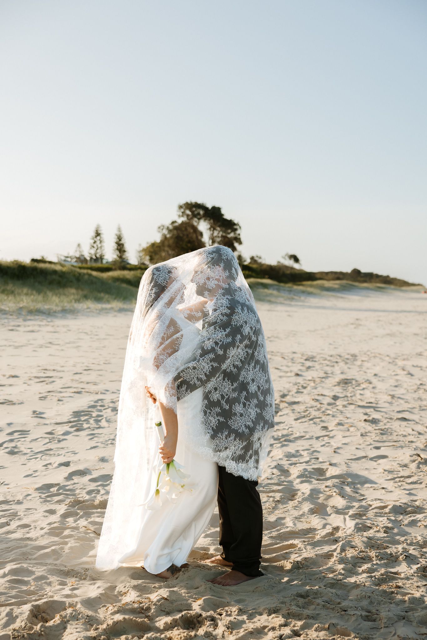 Bride and groom kissing under lace veil on sandy beach at golden hour with dunes in background.