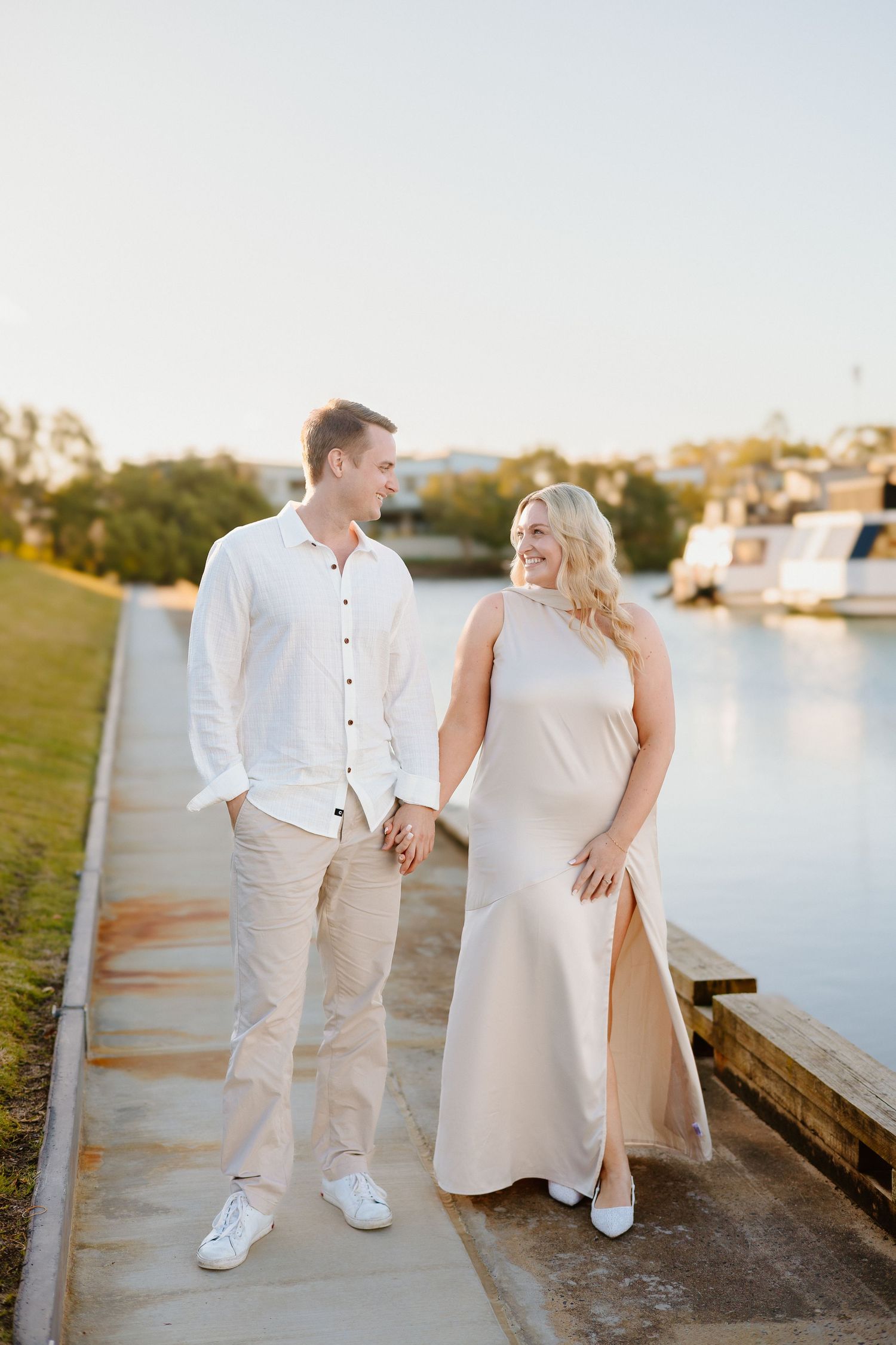 Couple holding hands and smiling by a waterway at golden hour, man in white shirt and woman in beige gown.