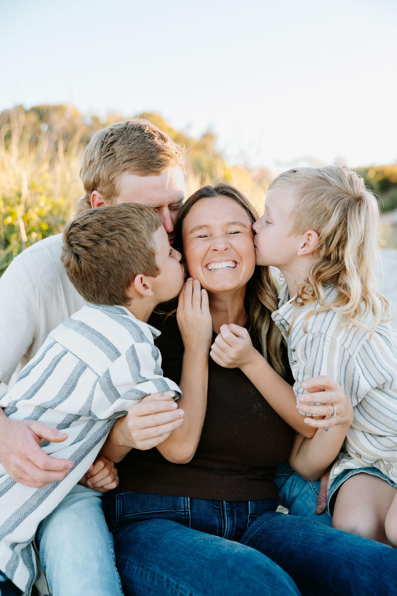 Family of four sharing a joyful moment outdoors, with kids kissing mom on cheeks while dad embraces them all.