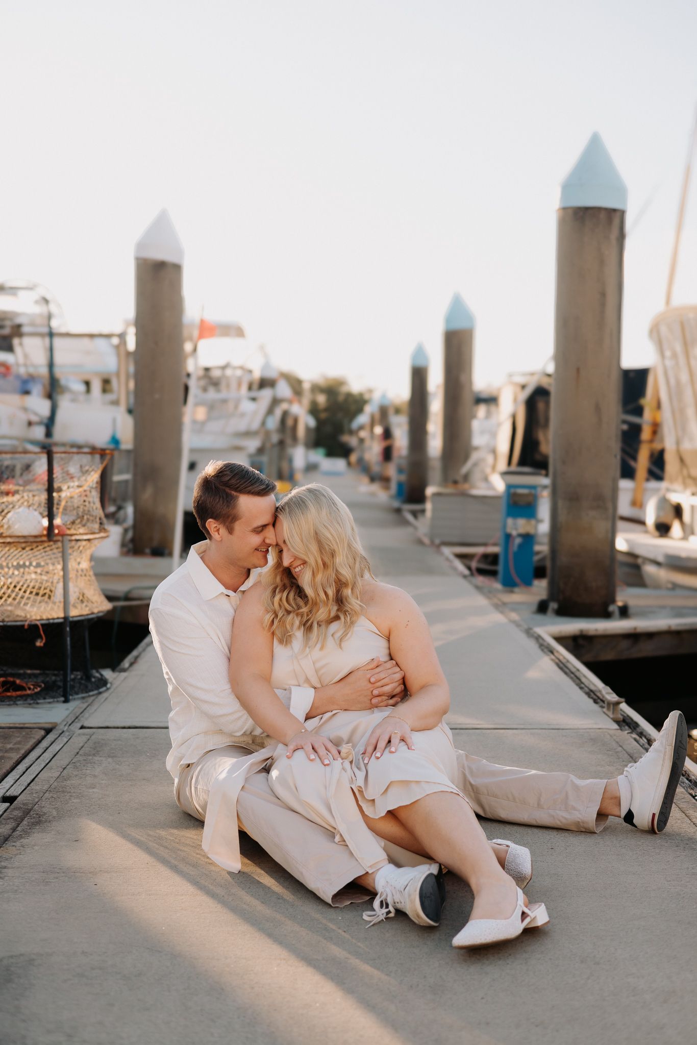 Couple sitting together on a marina dock at golden hour, sharing a romantic moment with boats in the background.