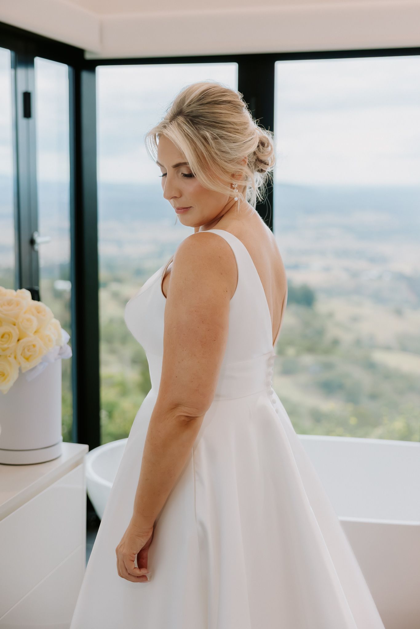Bride in elegant white A-line wedding dress poses by window with scenic valley views.