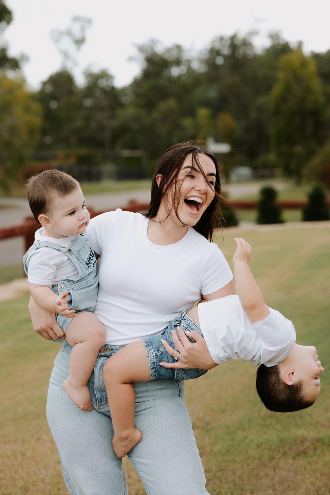 A laughing young woman holds two toddlers outdoors, one upright and one upside down, in a lush green park setting.