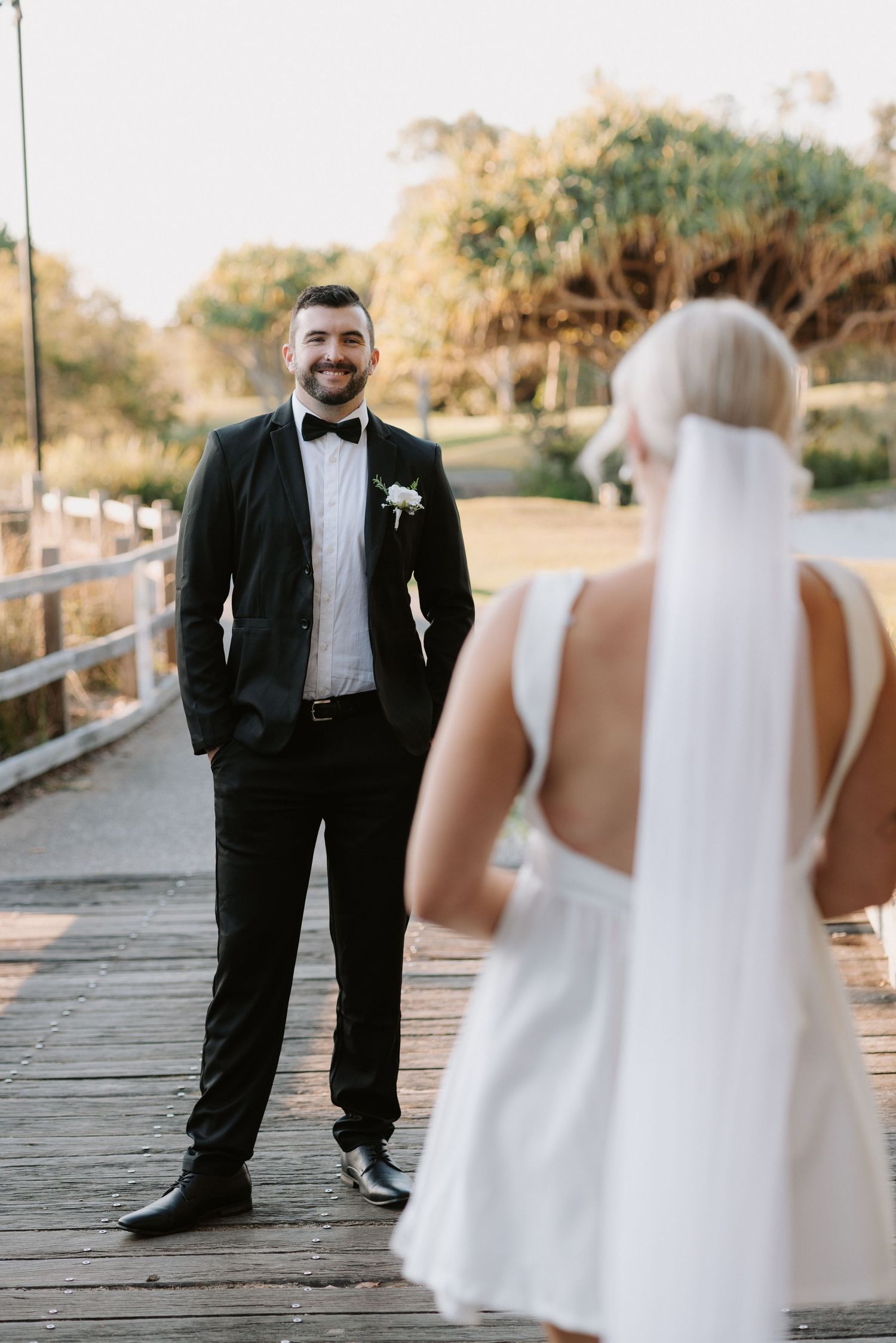 Smiling groom in black tuxedo stands on wooden bridge facing bride in white dress and veil during outdoor wedding.