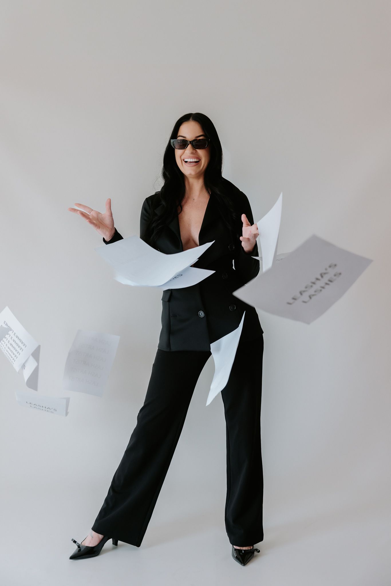 Woman in black suit and sunglasses tossing papers in the air, smiling confidently against a light background.
