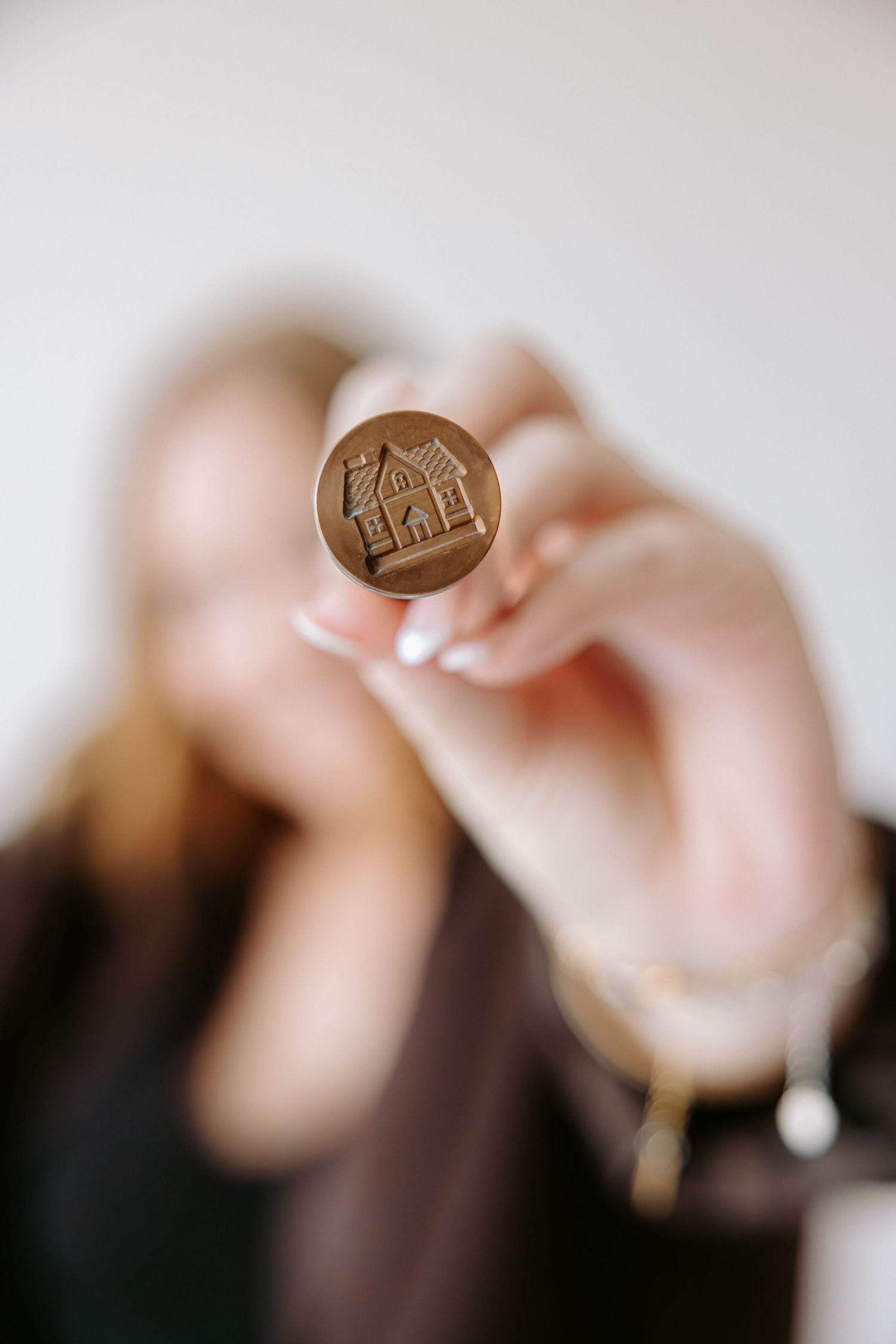A blond woman holds up a bronze wax seal stamp engraved with a house design toward the camera.