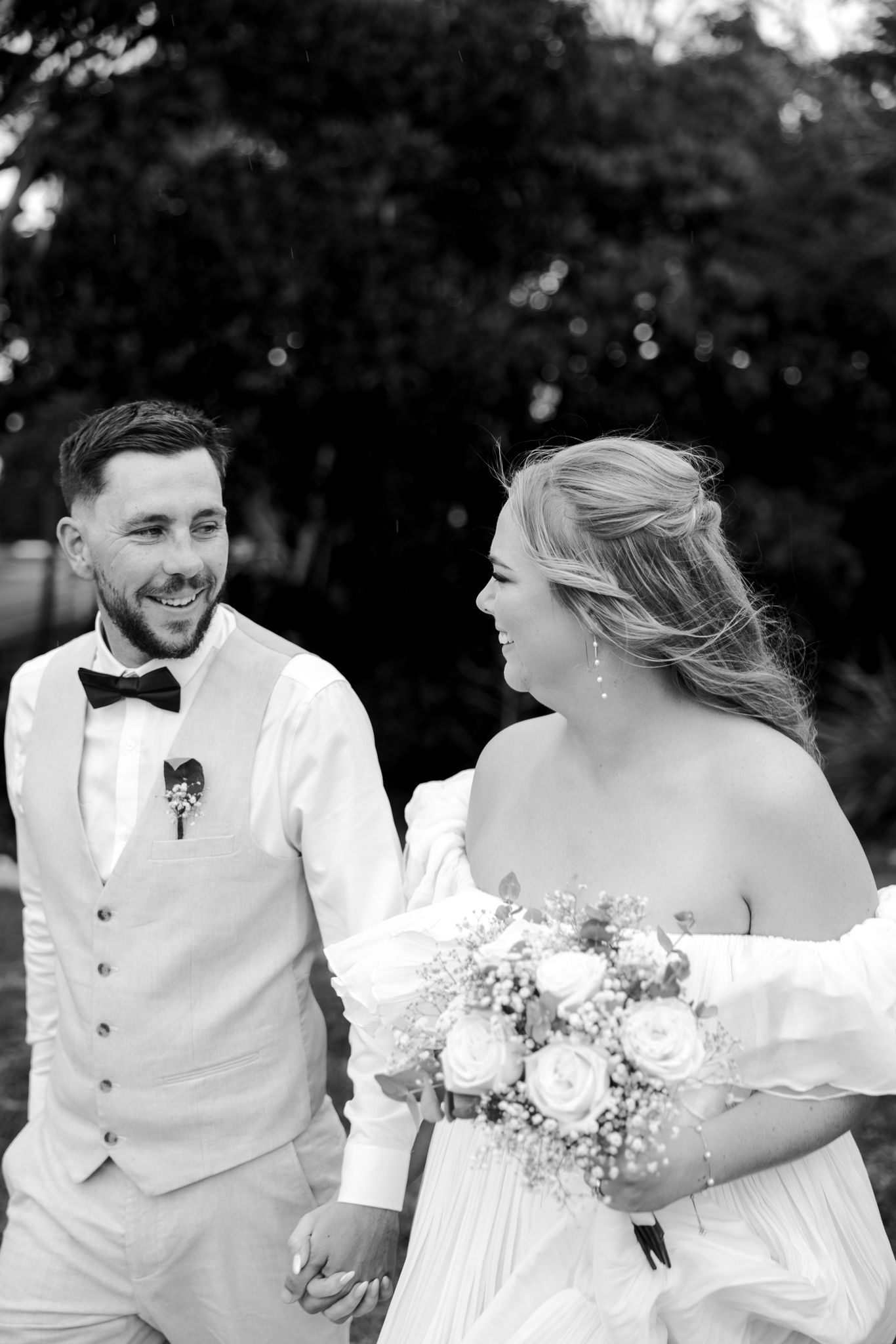 Black and white photo of smiling bride and groom holding hands outdoors, bride holding rose bouquet.