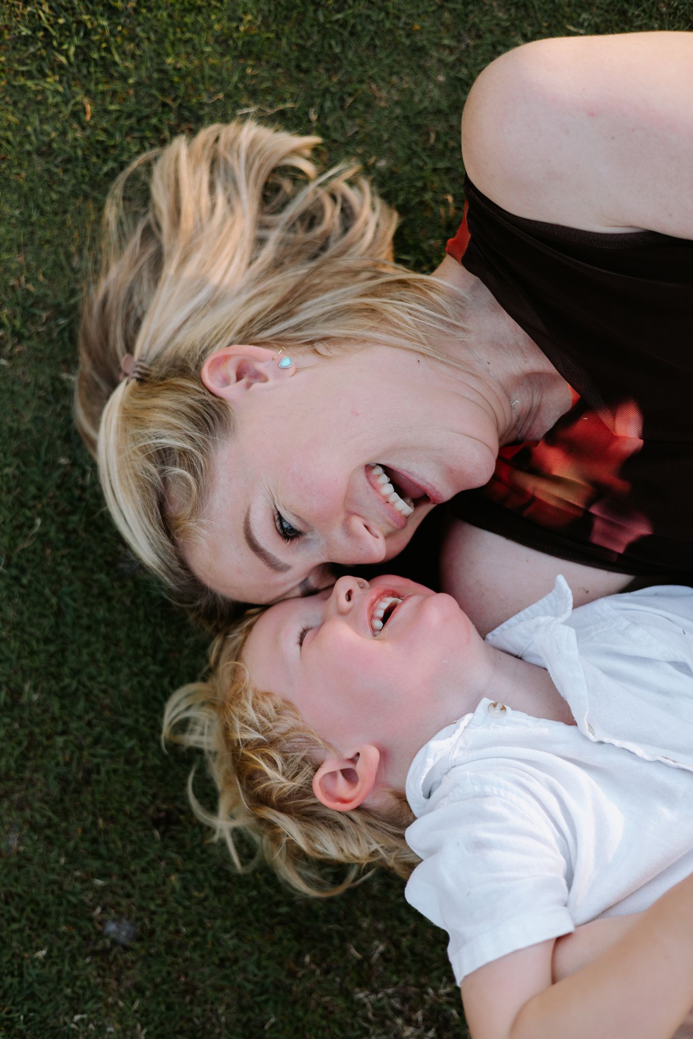 A joyful mother and toddler with blonde hair laughing together while lying on green grass outdoors.