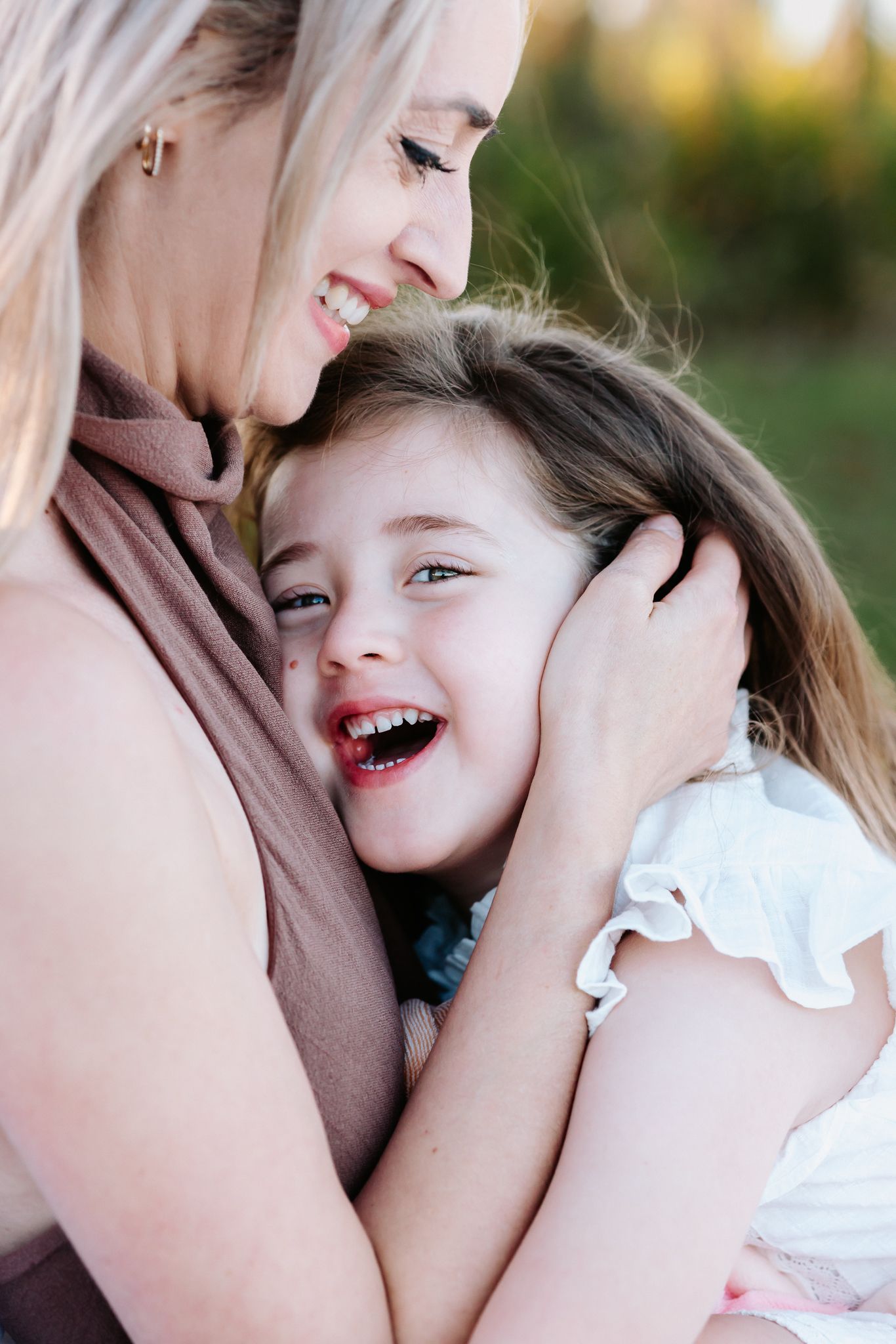 A smiling blonde mother tenderly embraces her laughing young daughter outdoors in warm natural light.