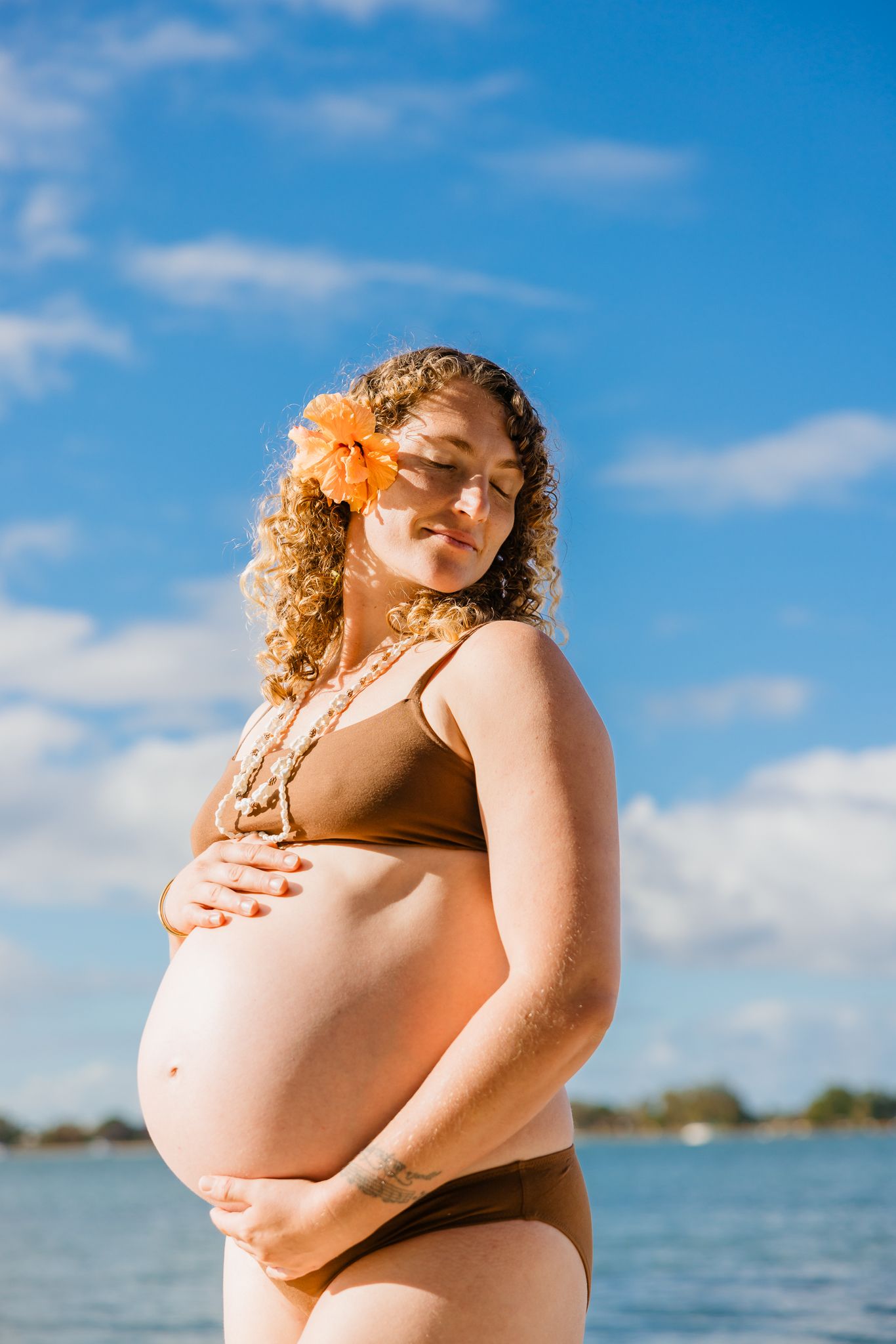 Pregnant woman in brown bikini with orange flower in curly hair, cradling her belly by the ocean under blue sky.