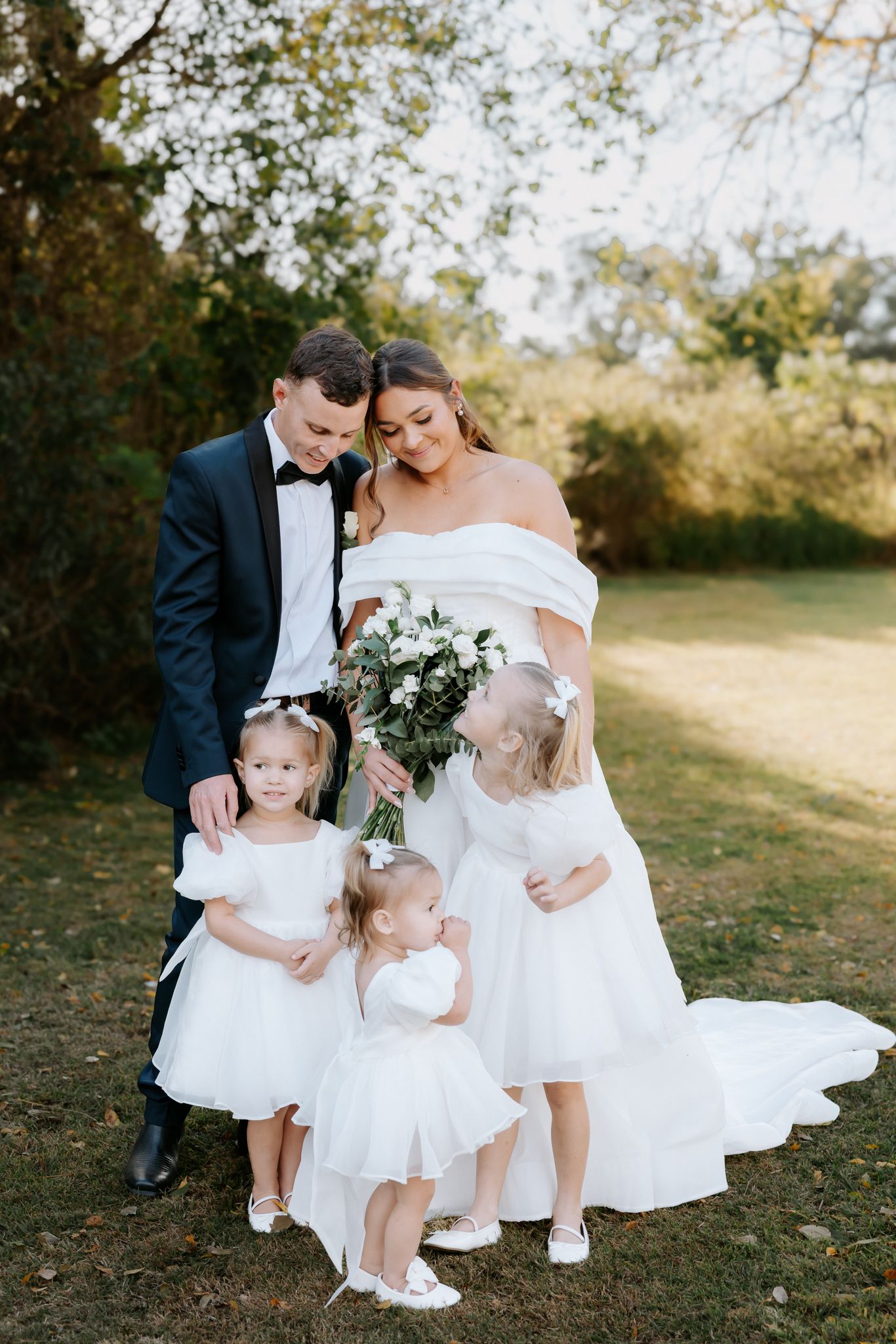 Bride and groom in formal wedding attire pose outdoors with three young flower girls in white dresses on a sunny day.