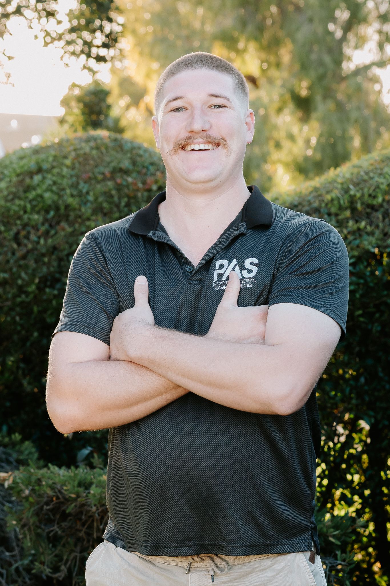Smiling man with mustache in dark PAS polo shirt, arms crossed, standing outdoors near green hedges.