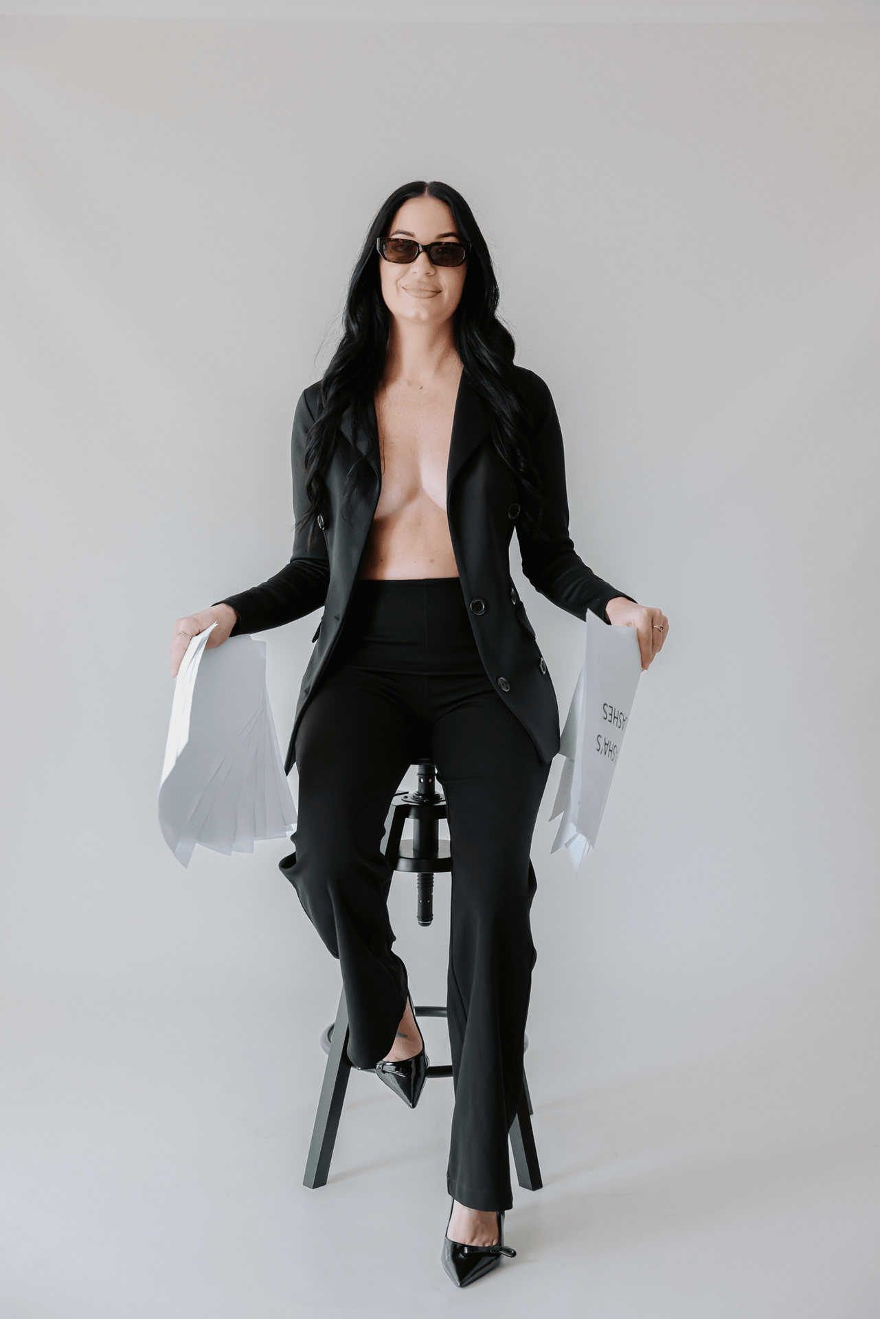 Woman in black blazer suit and sunglasses sitting on stool holding white bags against light background.