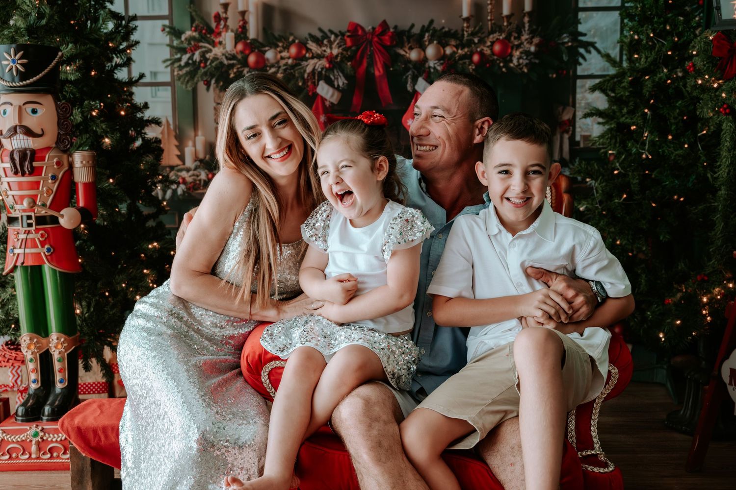 Family of four smiling together in front of Christmas trees and holiday decorations during a festive photo session.