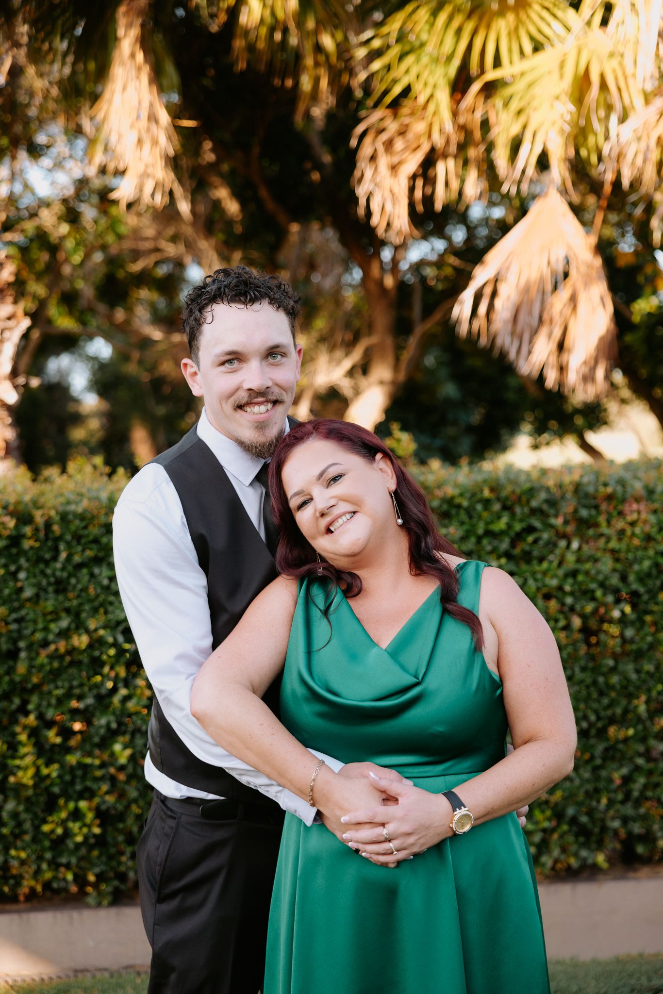 A smiling couple poses outdoors, man in vest embracing woman in elegant green satin dress, with palm trees in background.