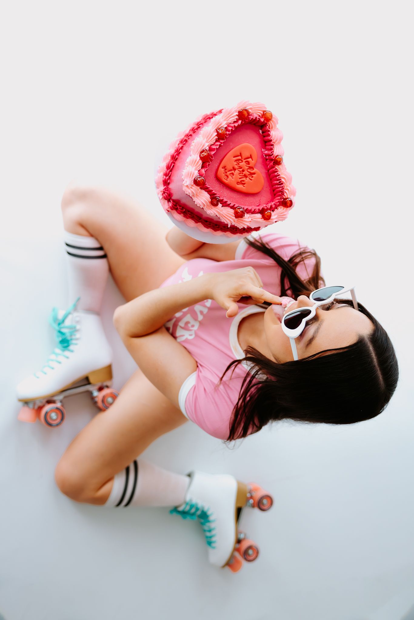 Woman in pink outfit and roller skates lying down holding a heart-shaped Valentine's Day cake with sunglasses.