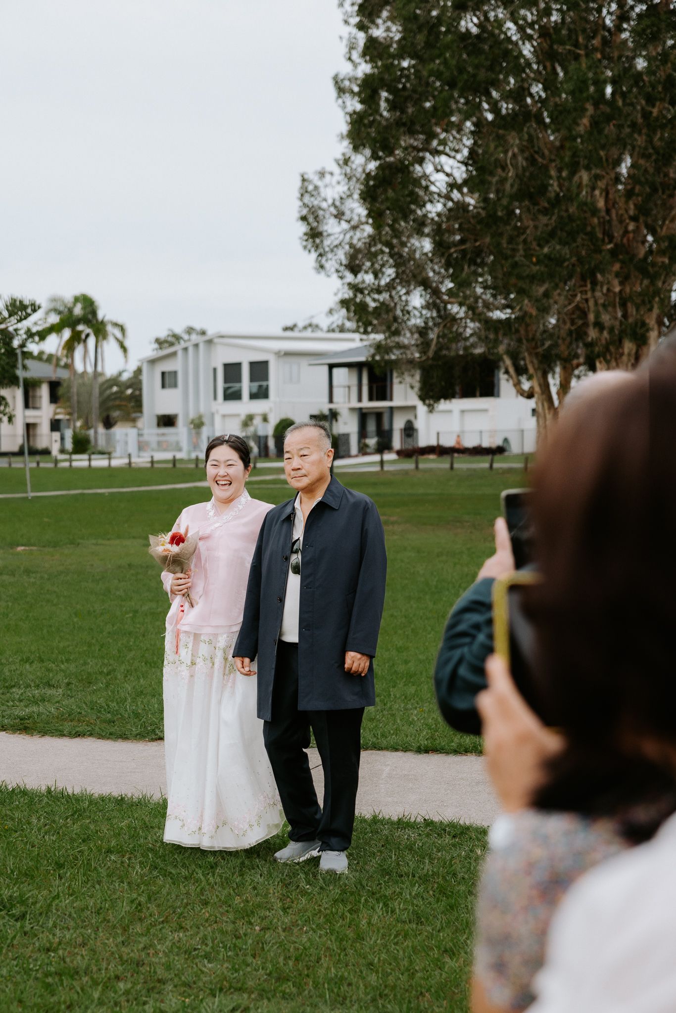 Couple posing for a photo on green lawn with white building and trees in background.