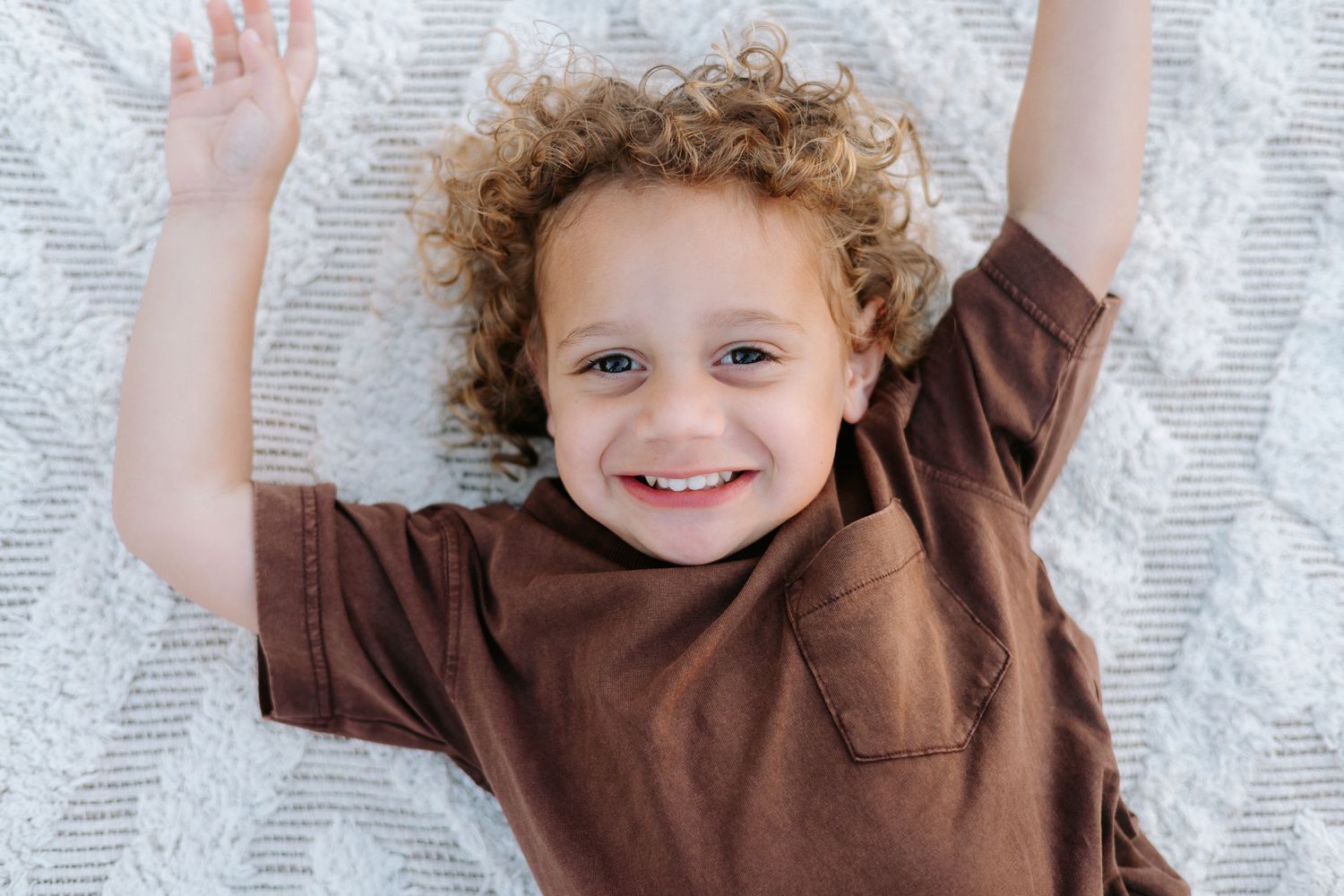 Happy toddler with curly hair lying on a white blanket, smiling with arms raised in joy.