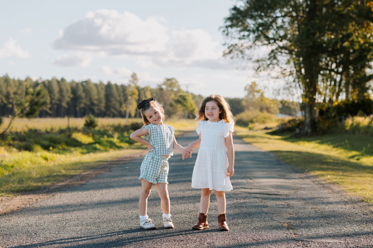 Two young girls in dresses hold hands on a rural road surrounded by trees and fields on a sunny day.