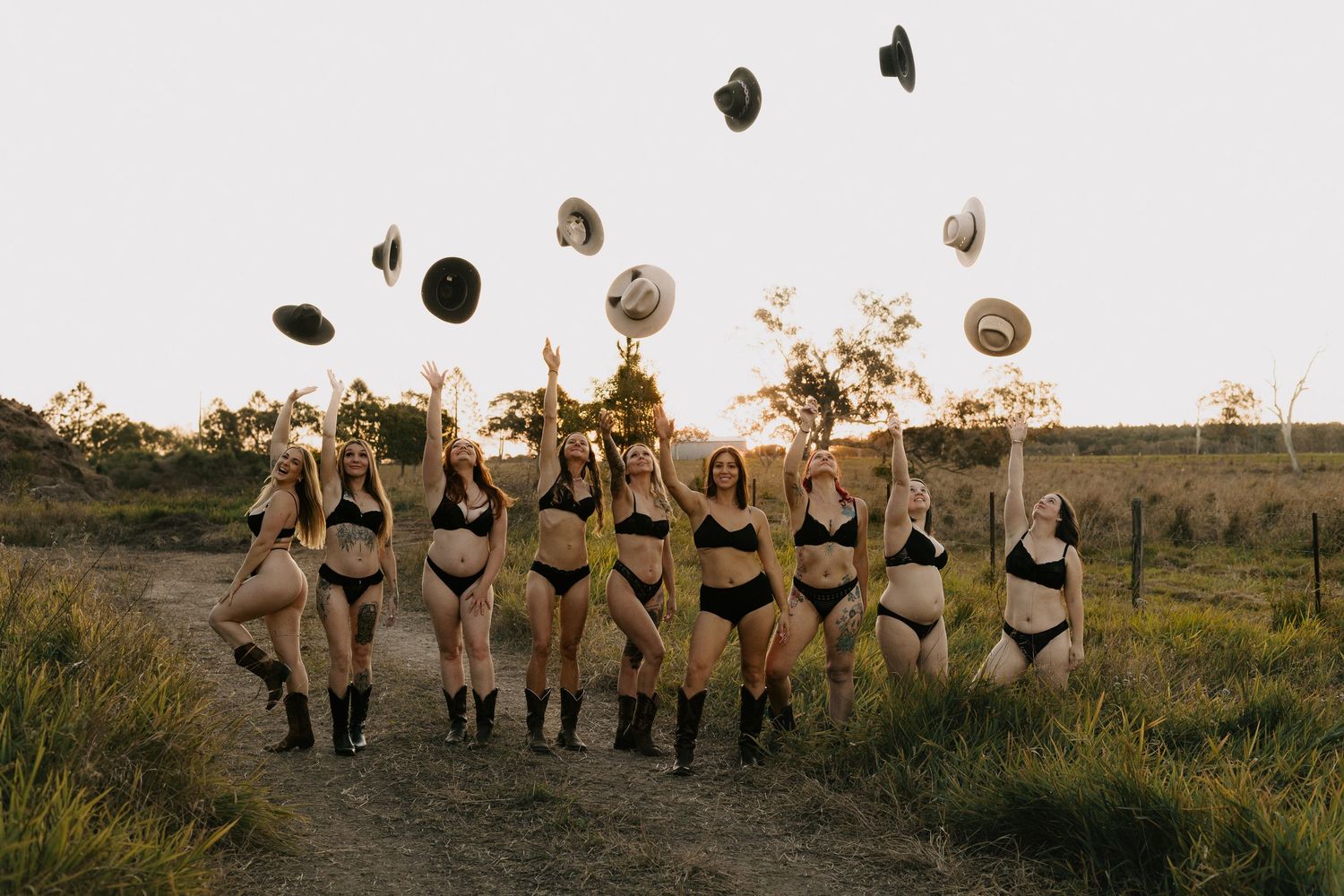 Group of women in black bikinis and cowboy boots tossing hats in the air on a rural dirt path at sunset.