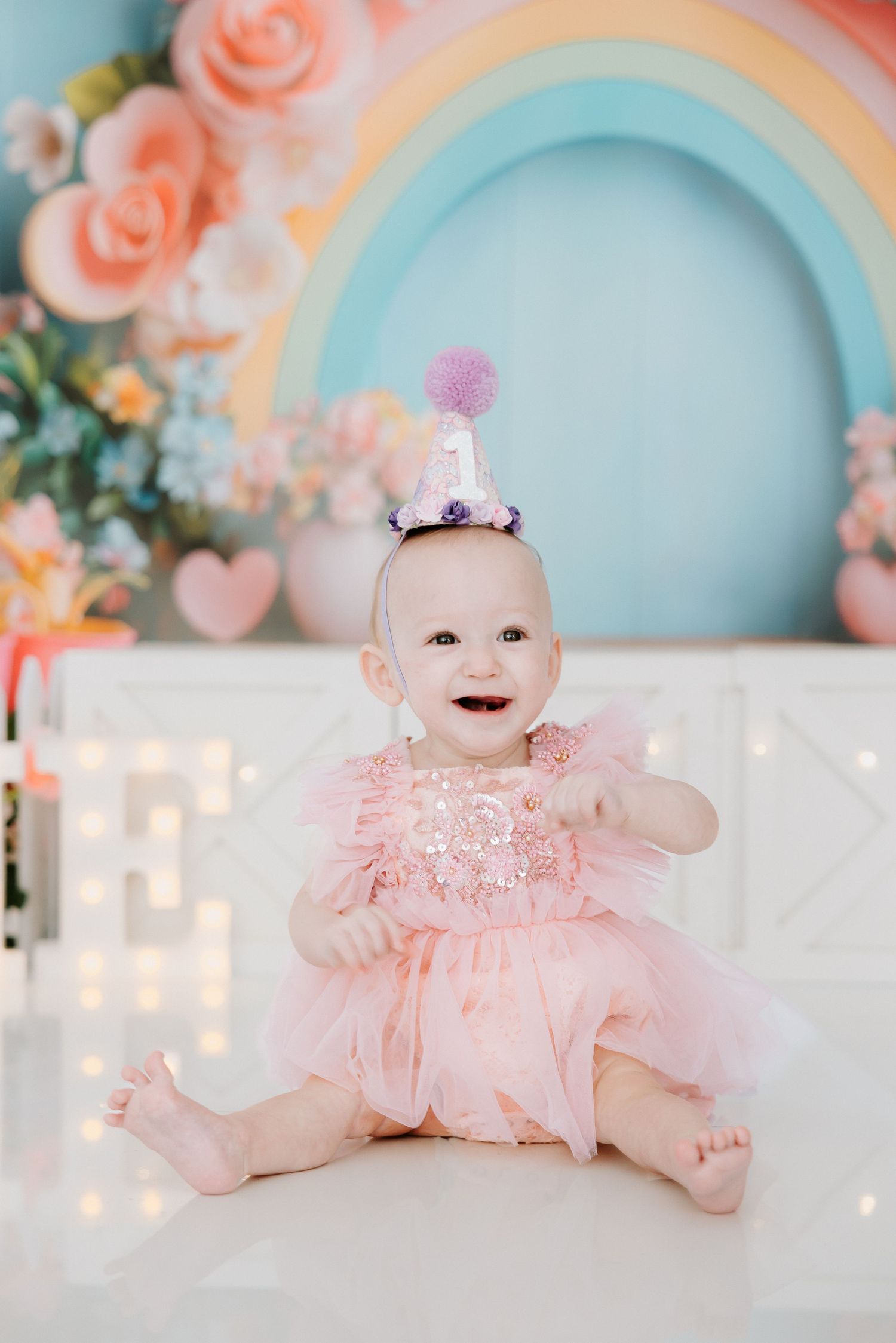 A smiling baby girl in a pink tutu dress and party hat sits in front of a colorful rainbow backdrop for her first birthday.