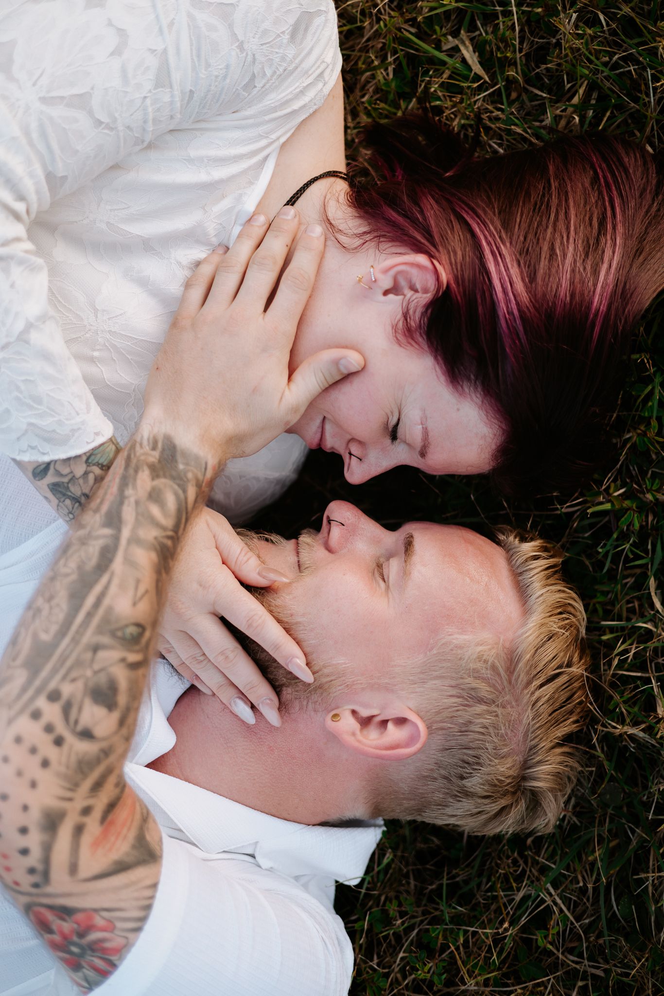 Couple lying on grass, woman with red hair and tattoos tenderly touching man's face in romantic moment.