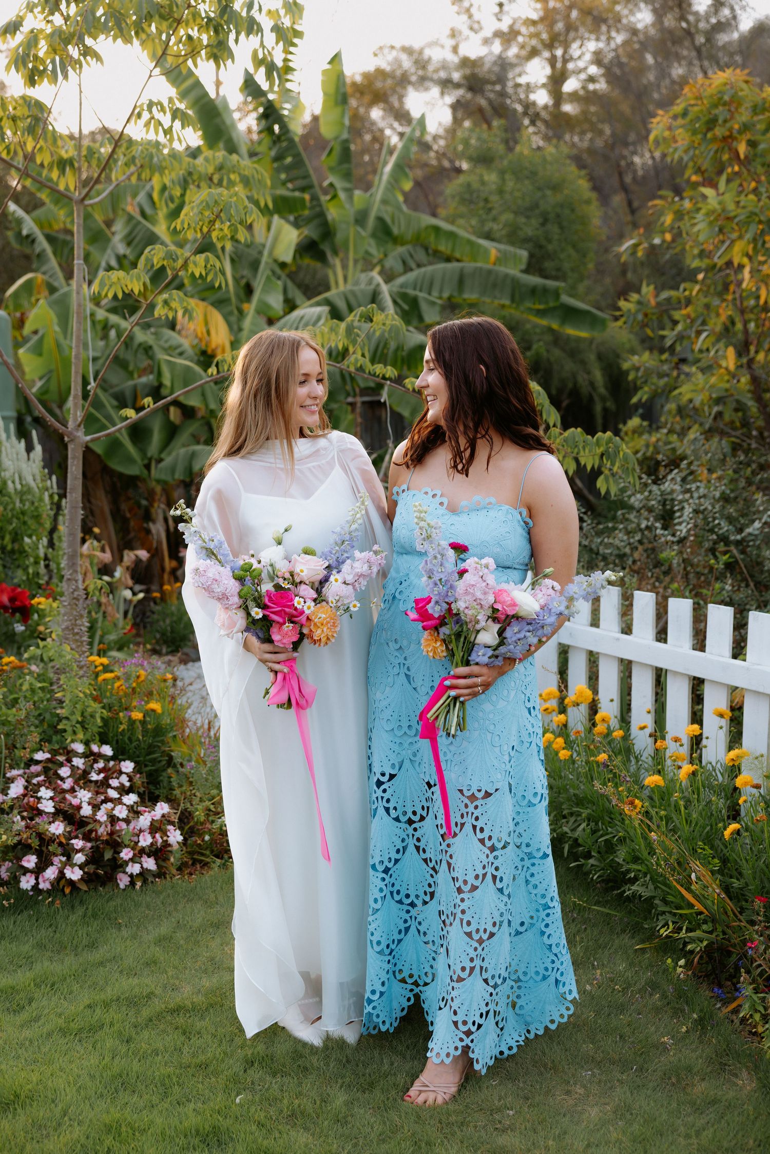 Two people in white and blue dresses walk through a garden with flowers.