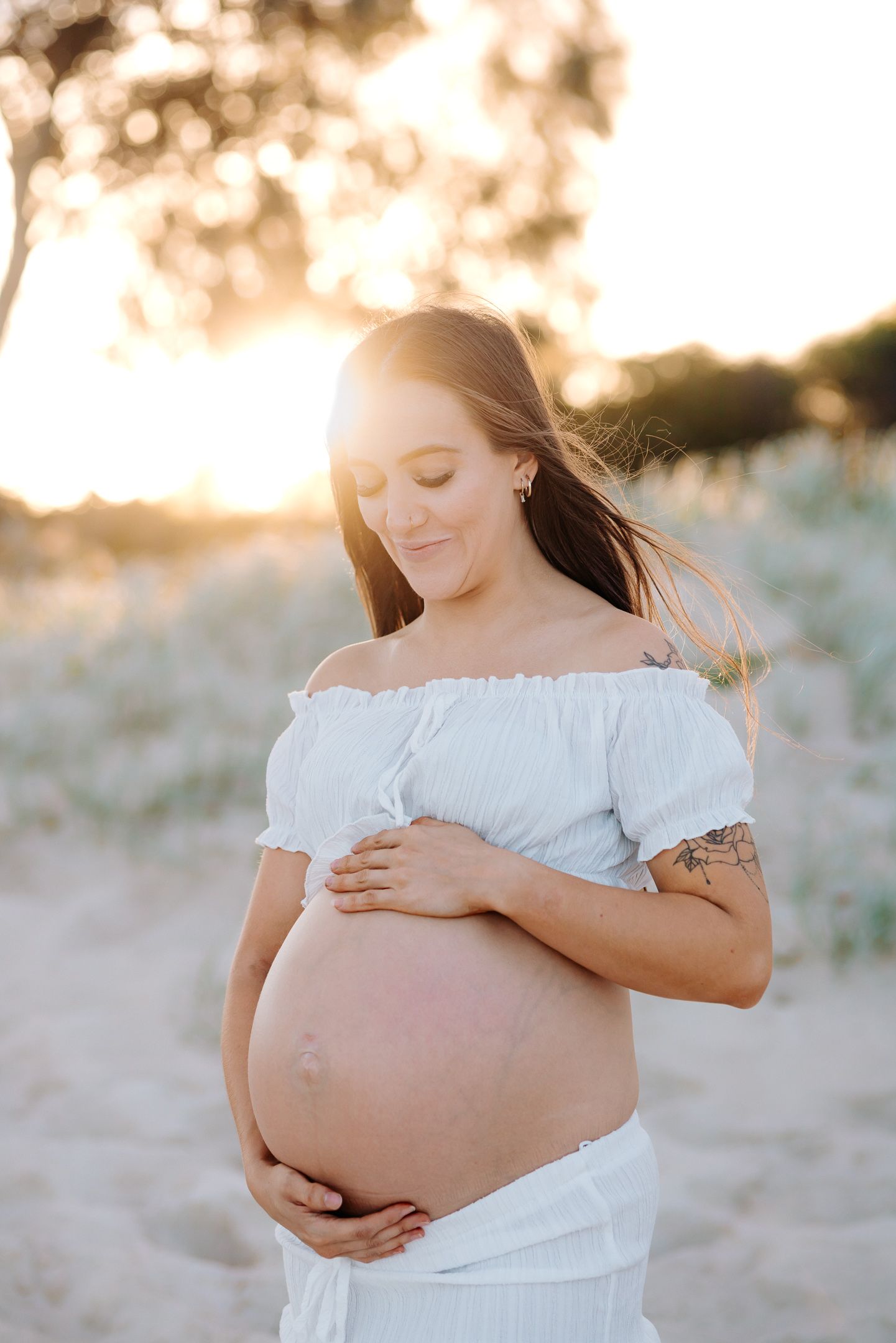 A maternity photo series shows a woman in a white off-shoulder dress cradling her baby bump on a sandy beach at sunset.