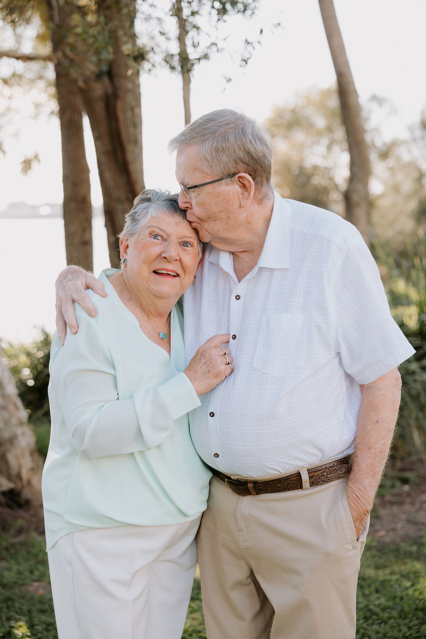 Elderly couple sharing a tender moment outdoors, man kissing womans head while embracing near trees.