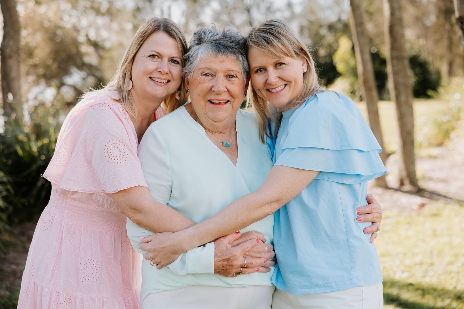 Three women smiling and hugging outdoors, two middle-aged women in pink and blue flank an elderly woman in white.