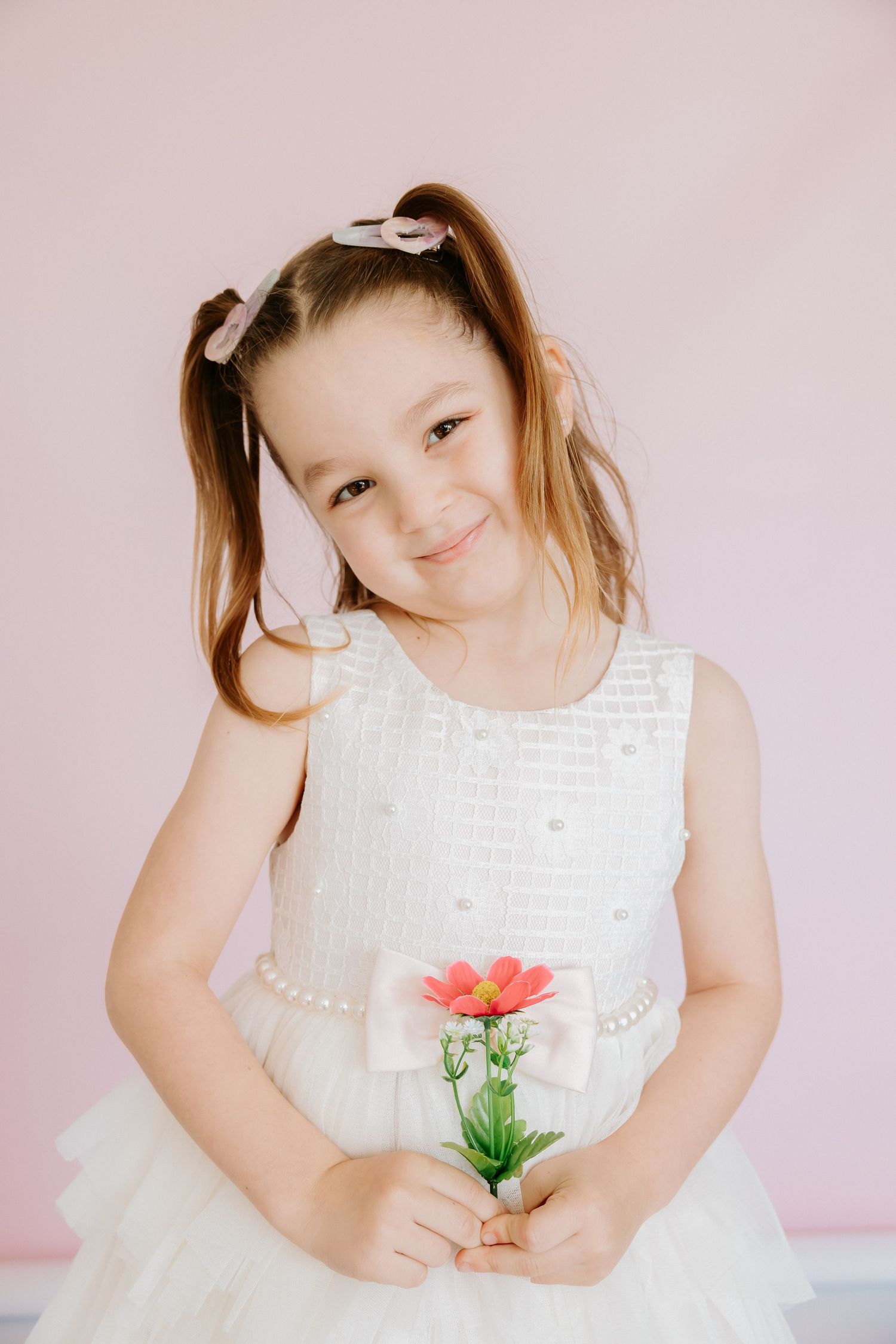 Young girl in white dress holding a pink flower, smiling against a soft pink background.