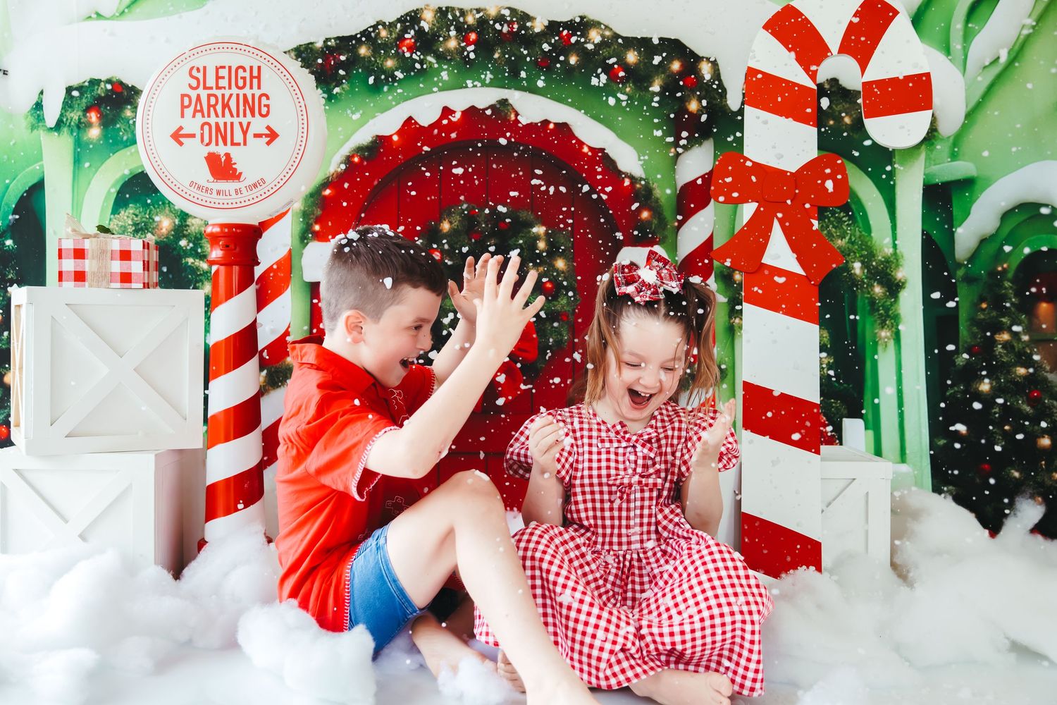 Two children in red outfits play together in a festive Christmas photo backdrop with candy canes and sleigh parking sign.