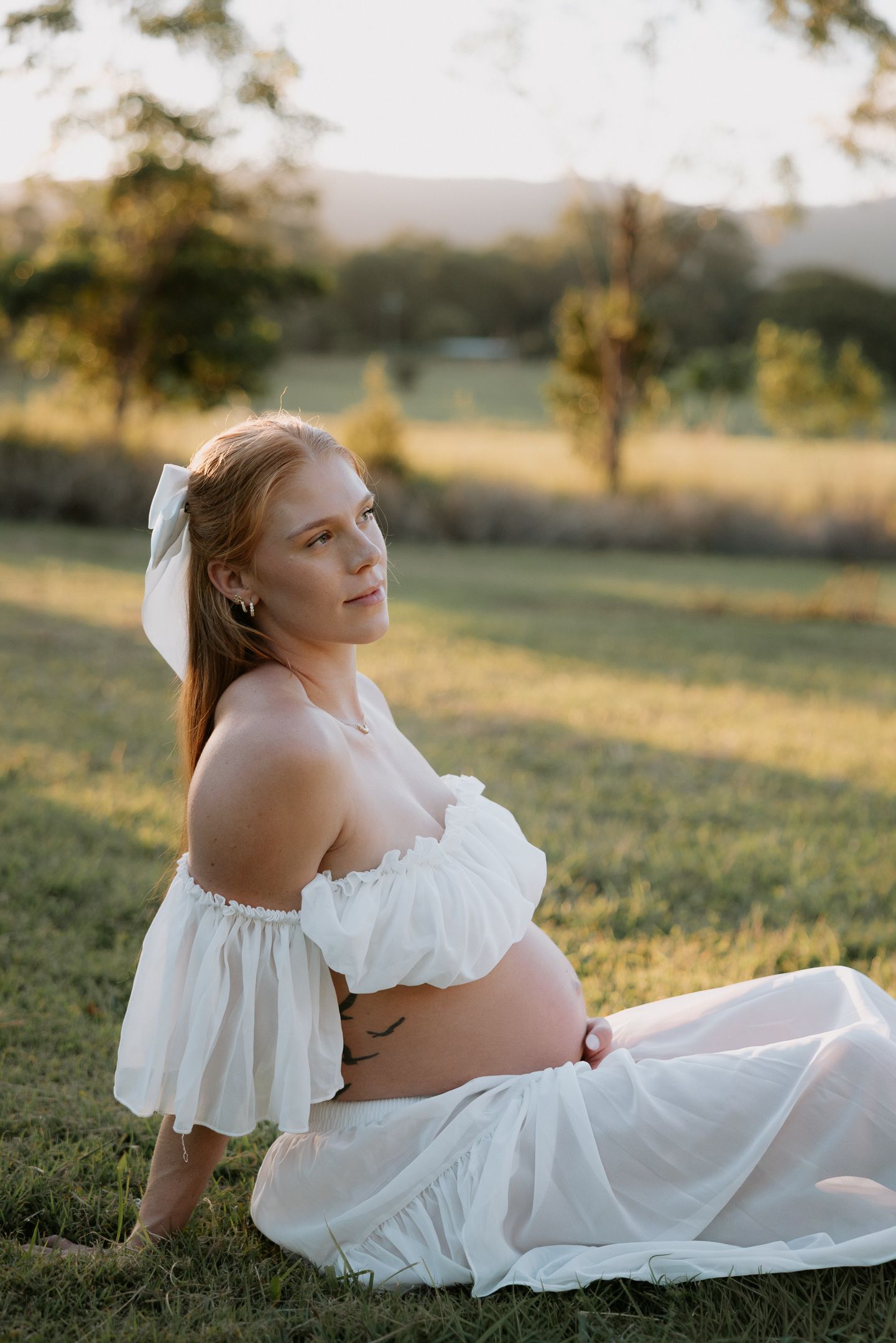 Pregnant woman in white off-shoulder dress sitting on grass during golden hour sunset maternity photoshoot.