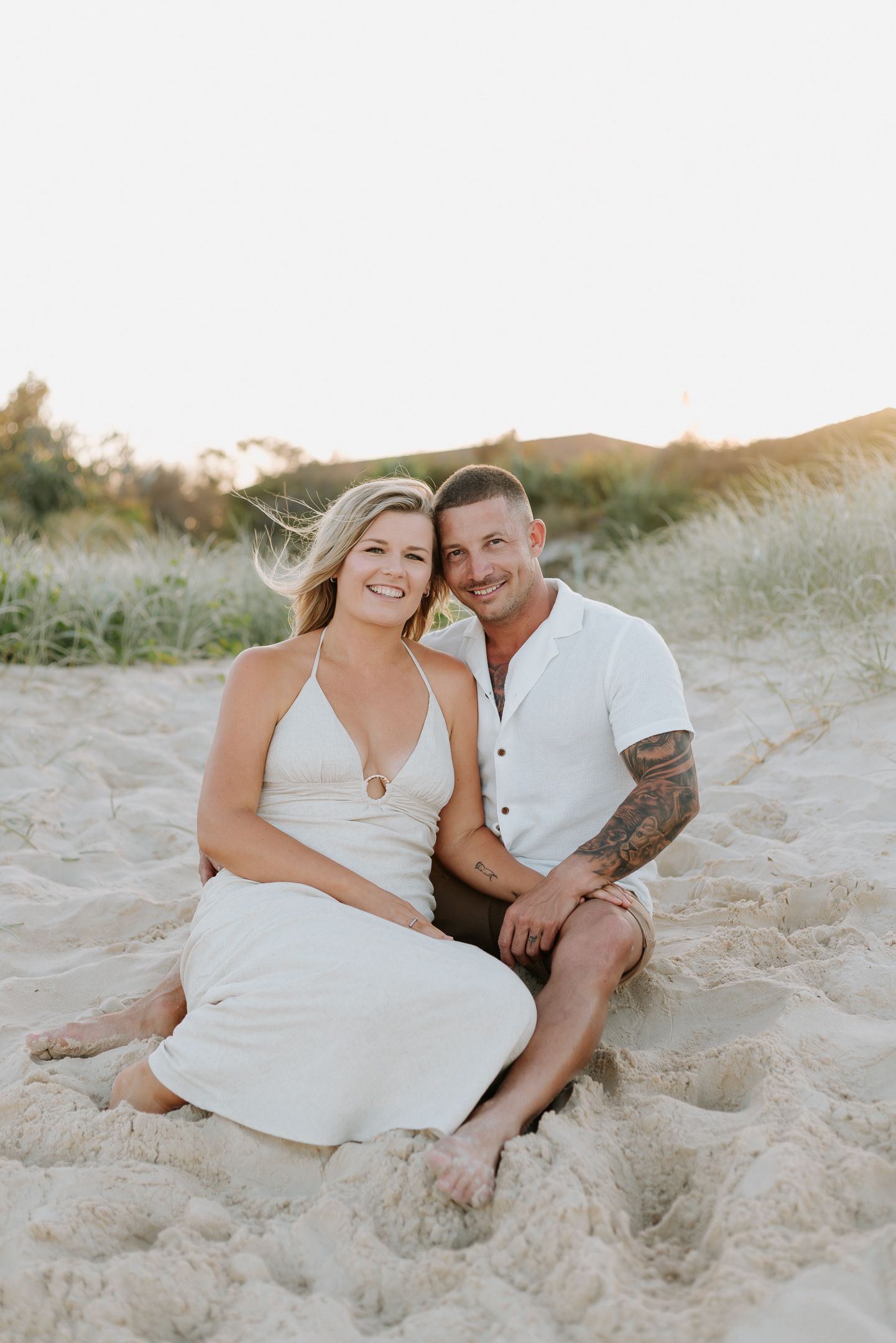 A smiling couple sits together on sandy beach dunes at golden hour, woman in white dress, man in white shirt with tattoos.