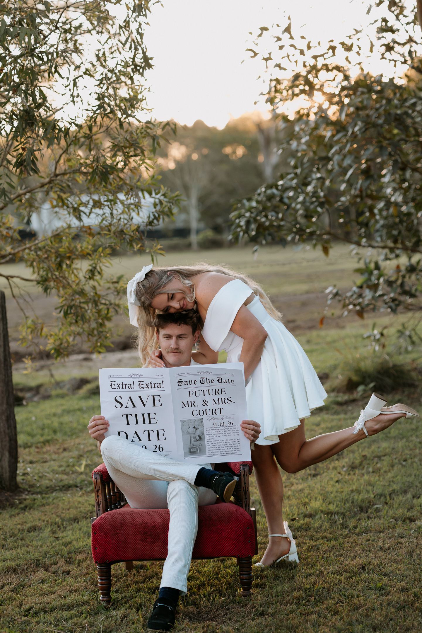 Couple poses with Save the Date newspaper sign outdoors, bride in white dress leaning over groom seated in red chair.