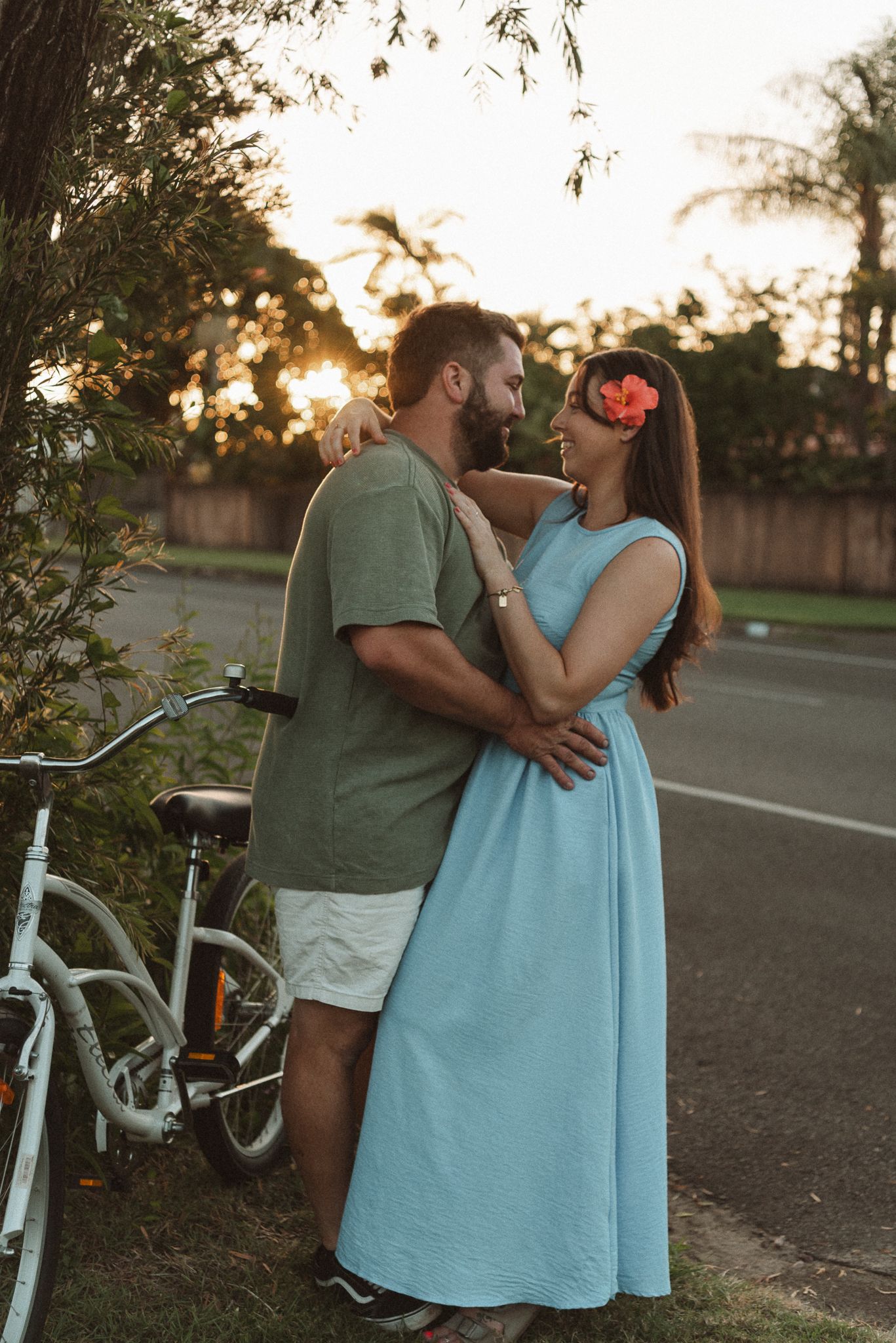 Couple embracing at sunset near a white bicycle, woman in blue dress with pink flower in hair.