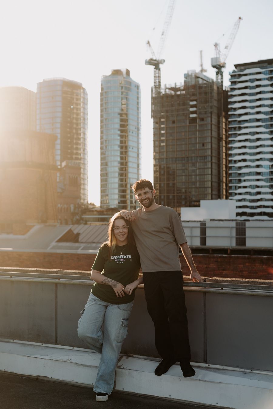 A couple poses on a rooftop with city skyscrapers and construction cranes in the background at golden hour.