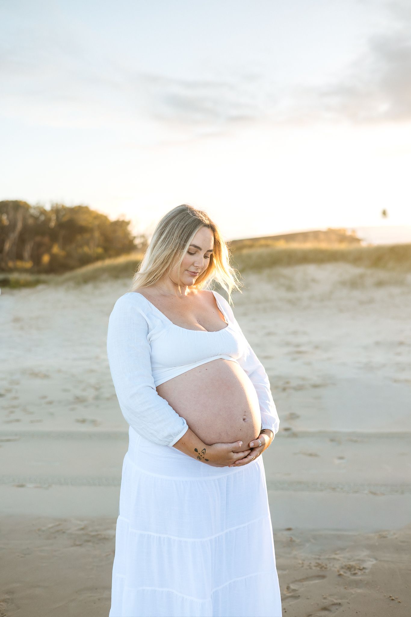Pregnant woman in white dress cradling baby bump on sandy beach at golden hour sunset.