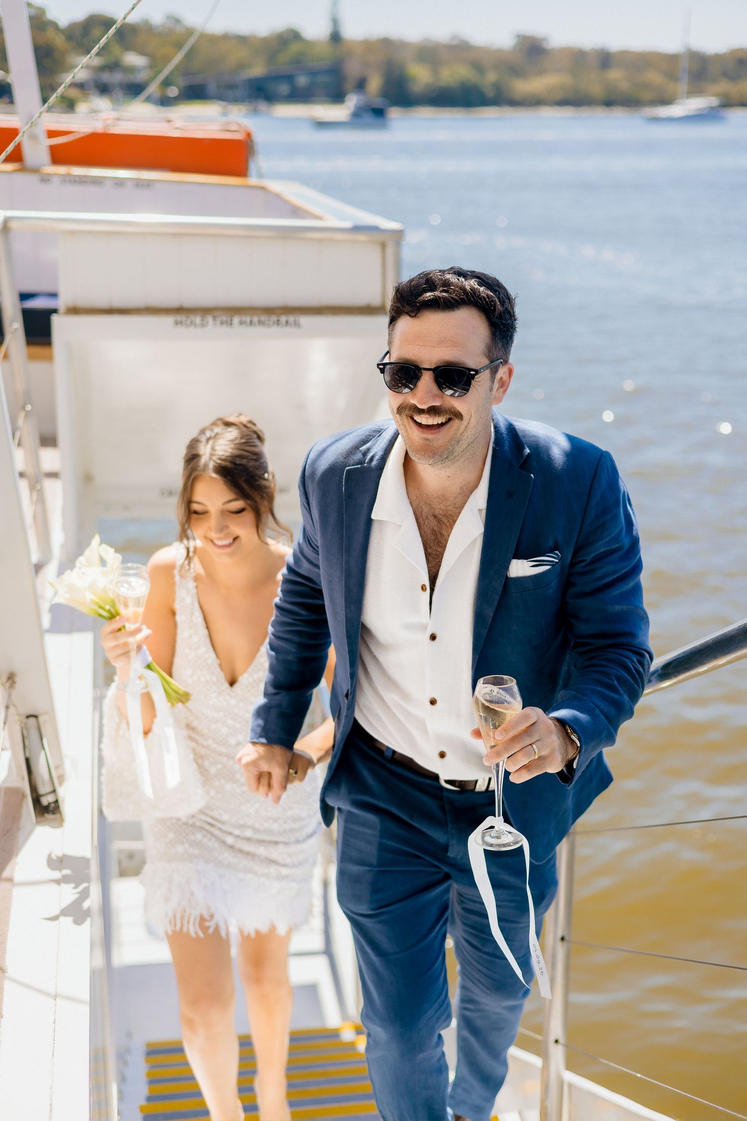 Bride and groom in navy suit holding champagne glasses walk off yacht on sunny wedding day.