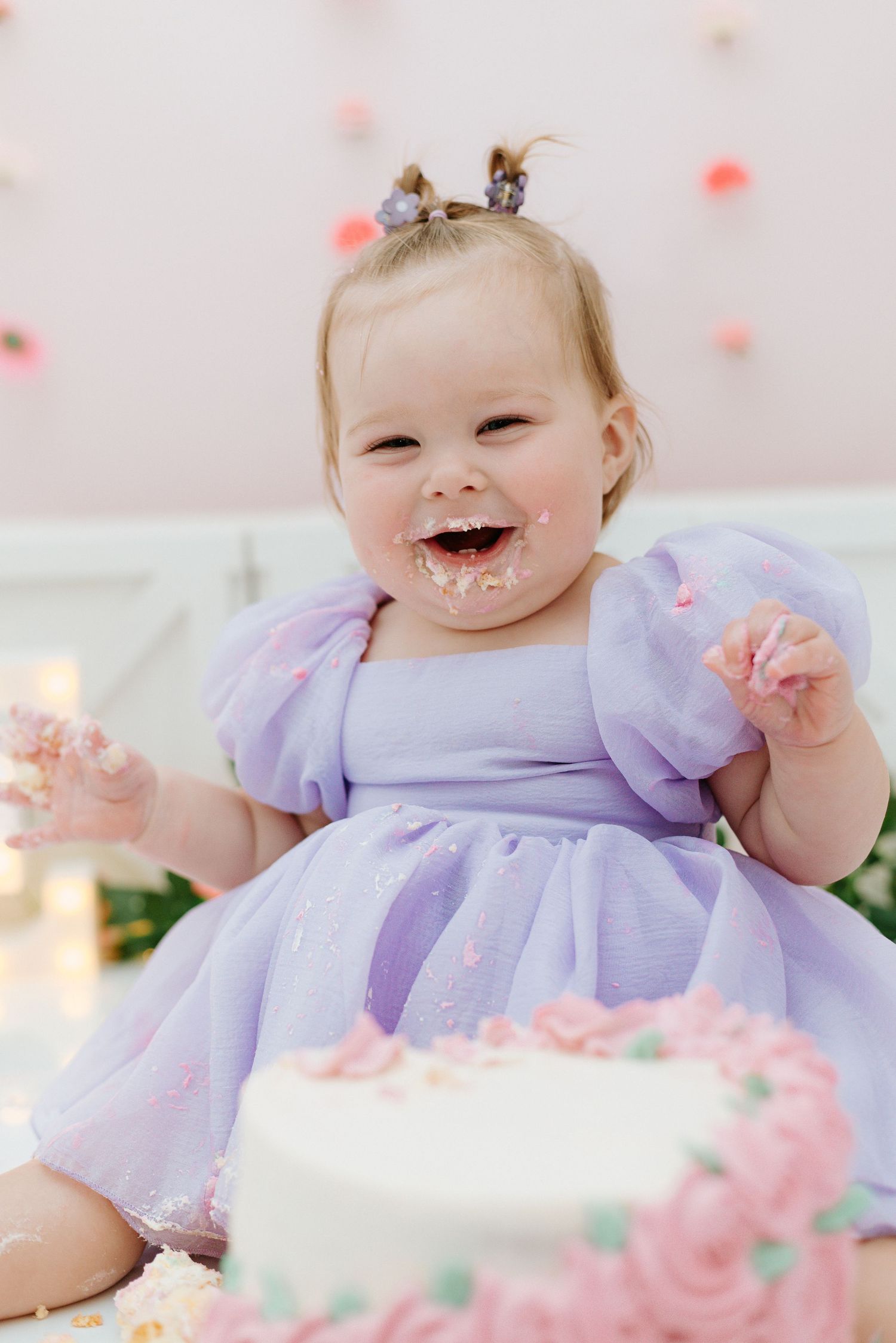 Baby in lavender dress smiles and plays with pink floral decorations during cake smash photo session.